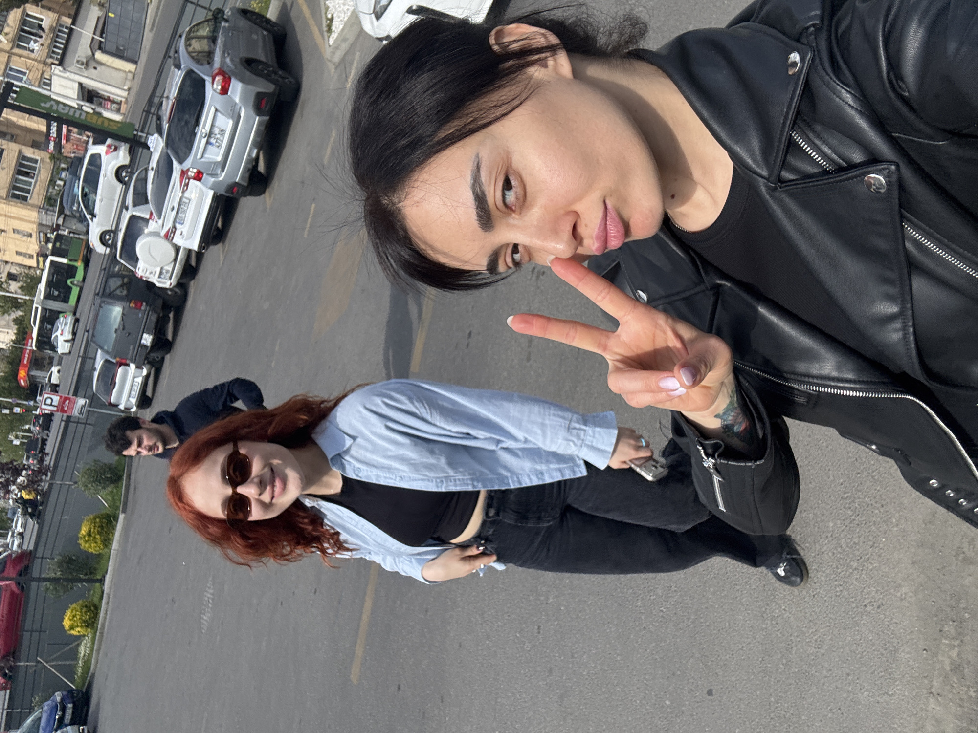 A tilted daytime selfie captures three individuals outdoors in what appears to be a parking area or street in Tbilisi, Georgia.
In the foreground, a woman with dark hair, wearing a black leather jacket and a black top, takes the selfie. She has defined eyebrows and lips, and is making a peace sign with her right hand, showcasing nails with light polish. A small black bag is visible on her left arm, and she has a slight smirk.
Behind her, to her left, stands a woman with reddish-brown hair, wearing sunglasses, a light blue unbuttoned long-sleeve shirt over a black crop top, and black jeans. She is holding a silver-colored mobile phone in her left hand and is smiling towards the camera. Further back and slightly to the right of the red-haired woman, a man with dark hair, wearing a dark blue top, is partially visible, looking towards the camera.
The background shows a line of parked cars along a street. Vehicles include a silver SUV with the Georgian license plate "NE-677-IN", a white SUV with a spare tire mounted on the back (license plate "GE 507 HI"), and other cars. Buildings line the street, with a green "Subway" sign visible on one building. A white "P" parking sign is also visible. The scene is brightly lit by natural sunlight, suggesting a clear day.