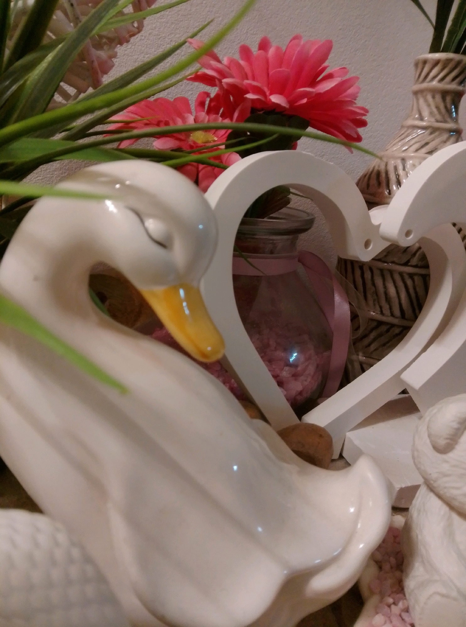 The image shows a close-up, indoor still life arrangement. The primary subject is a white ceramic swan with its eyes closed and its beak pointed slightly downwards. To the left and behind the swan, there are green artificial plant leaves and pink artificial flowers, some of which are in focus. To the right of the swan, a white heart-shaped decorative object is visible, with a glass jar containing pink decorative stones and a pink ribbon inside. Behind the heart, a beige ceramic vase with a textured design is present. The background consists of a textured off-white wall. There is no indication of the city of Wedel, Germany, or any other location. The scene is static and appears to be a decorative display.