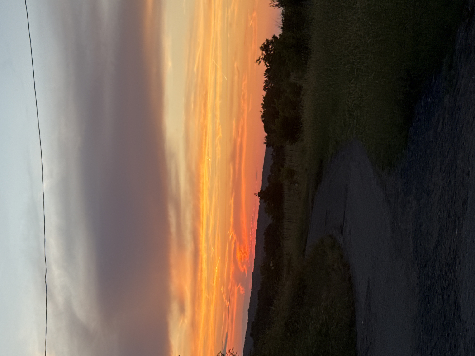 The image depicts a sunset over a rural landscape. The sky is filled with vibrant hues of orange, yellow, and pink, with streaks of darker clouds creating a dramatic effect. A single thin wire stretches horizontally across the upper portion of the sky. In the foreground, a gravel path curves through grassy fields and sparse vegetation. Trees and rolling hills form the horizon line, silhouetted against the colorful sky. The scene appears to be tranquil and natural, with no signs of urban development or immediate human presence. The location is identified as Krushevets, Bulgaria.
