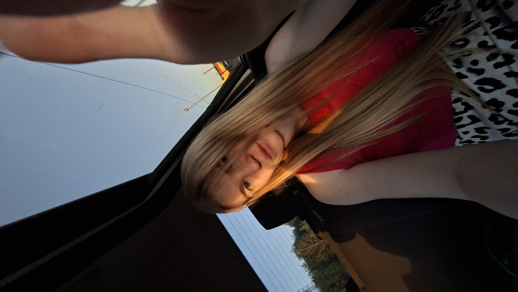 A selfie shows a blonde woman in a car. She's wearing a red top under a patterned shirt. The sky is visible through the car window, and trees are seen in the distance. The photograph was likely taken in the evening. A street light is visible through the car window. Without more context, the specific location in Chaikovsky, Russia, cannot be determined.