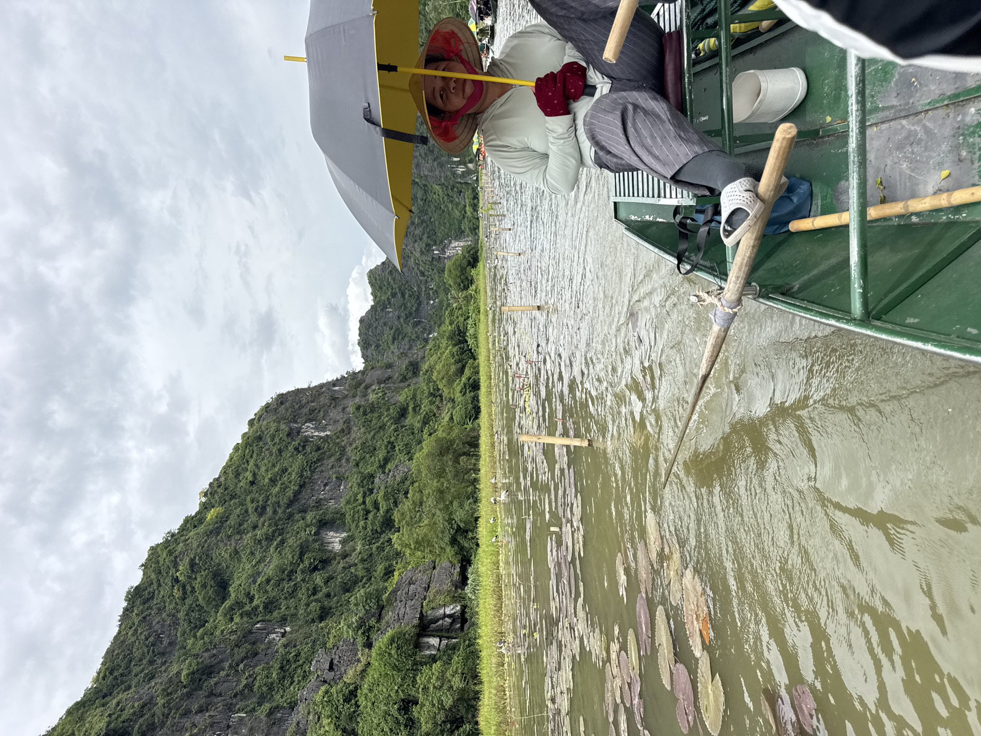 A person wearing a conical hat and holding a yellow and gray umbrella is seated in a green boat, looking towards the camera. They are wearing a light blue hooded jacket and dark striped pants. The boat is on a body of water with lily pads and wooden poles sticking out of the water. In the background, there is a lush green mountain and a cloudy sky. Several other boats and people can be seen in the distance. The scene appears to be a scenic waterway in Nam Hoa Lư Ward, Vietnam, possibly a tour or transportation route.