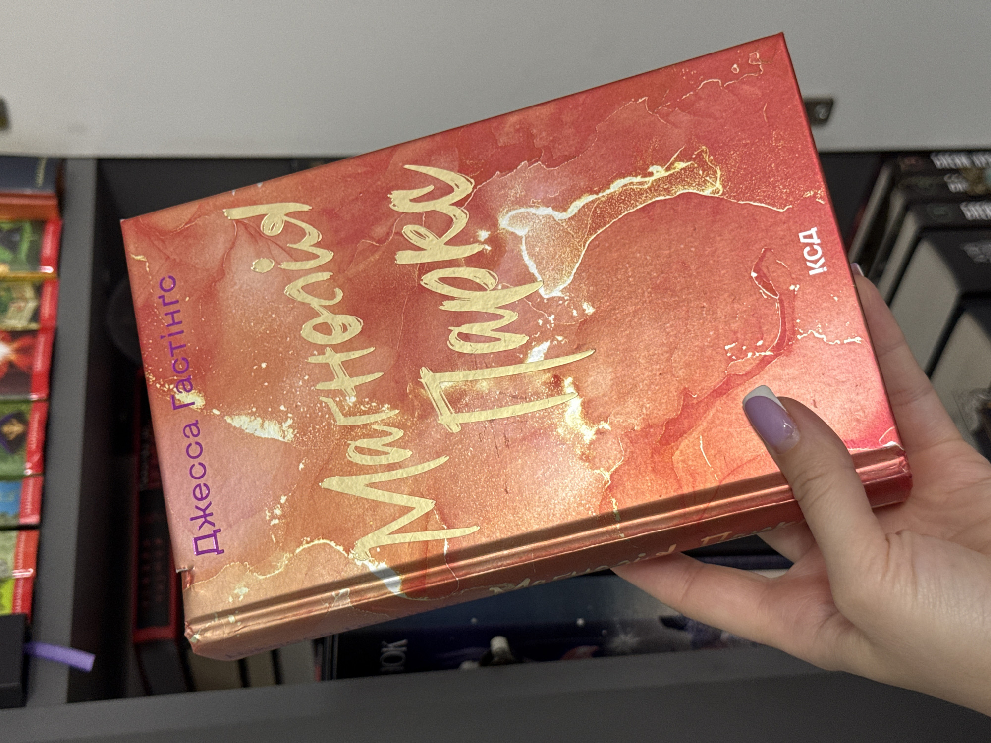 The image shows a close-up of a book with a vibrant, abstract red and gold cover, held by a person's hand. The book is angled, with its spine facing the viewer and its cover tilted upwards. The person's hand, with a lavender and white-tipped fingernail, gently supports the book from underneath, indicating care or examination. The background suggests a shelf or storage area, with other books partially visible on either side. The overall scene is well-lit, with a soft focus on the background elements, drawing attention to the book. The time of day is not discernible, but the indoor setting suggests it could be anytime. The emotion conveyed is one of quiet contemplation or appreciation for the book.
Visible text on the book includes:
- "Джесса Растінгс" (Dzhessа Rastings) in purple lettering along the left side of the cover.
- A title in stylized gold lettering that appears to read "Маргарита Паркер" (Margarita Parker).
- "КСД" (KSD) in small white lettering on the right side of the cover.
- The word "BOOK" is visible on the spine of a book in the background.
The location context of Łódź, Poland, does not provide specific visual cues within the image itself, but it informs the cultural context of the visible text.