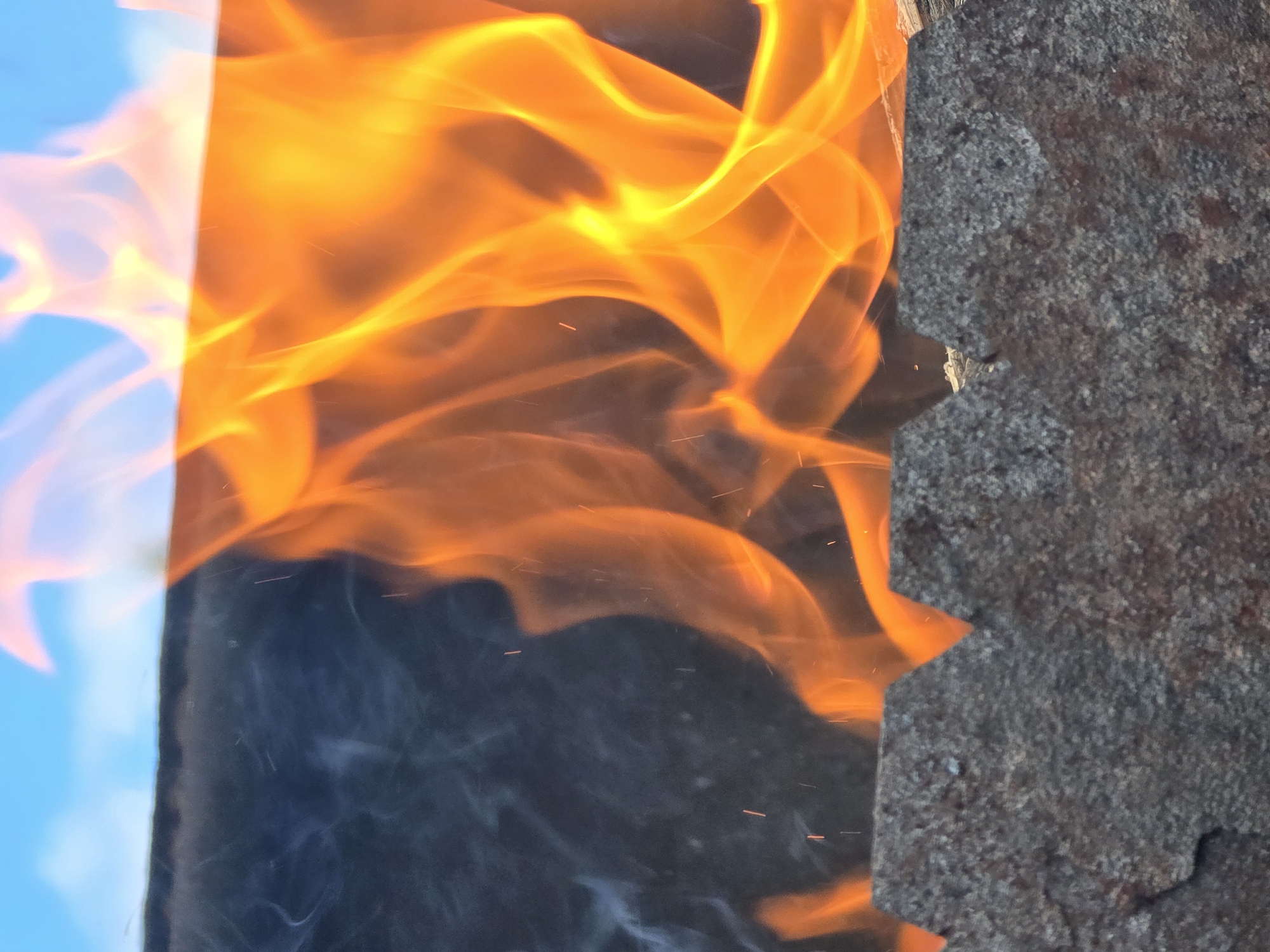This image is a close-up, low-angle shot of a fire burning in a metal grate. The flames are bright orange and yellow, with some wisps of white smoke to the left. The metal grate appears to be made of rough, dark-colored material with a jagged edge at the top. In the background, a clear blue sky is visible. There are no people or animals in the image. The scene is likely outdoors.