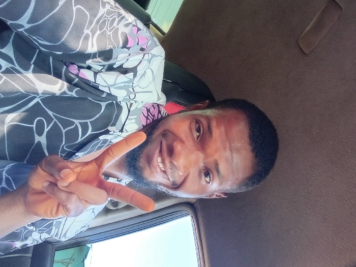 A man with dark hair and a beard smiles, holding up a peace sign in the passenger seat of a car. He is wearing a patterned shirt with dark, grey, and white colors, along with touches of pink. The interior of the car is brown. A window is visible, showing a bright blue sky and the top of a green structure.