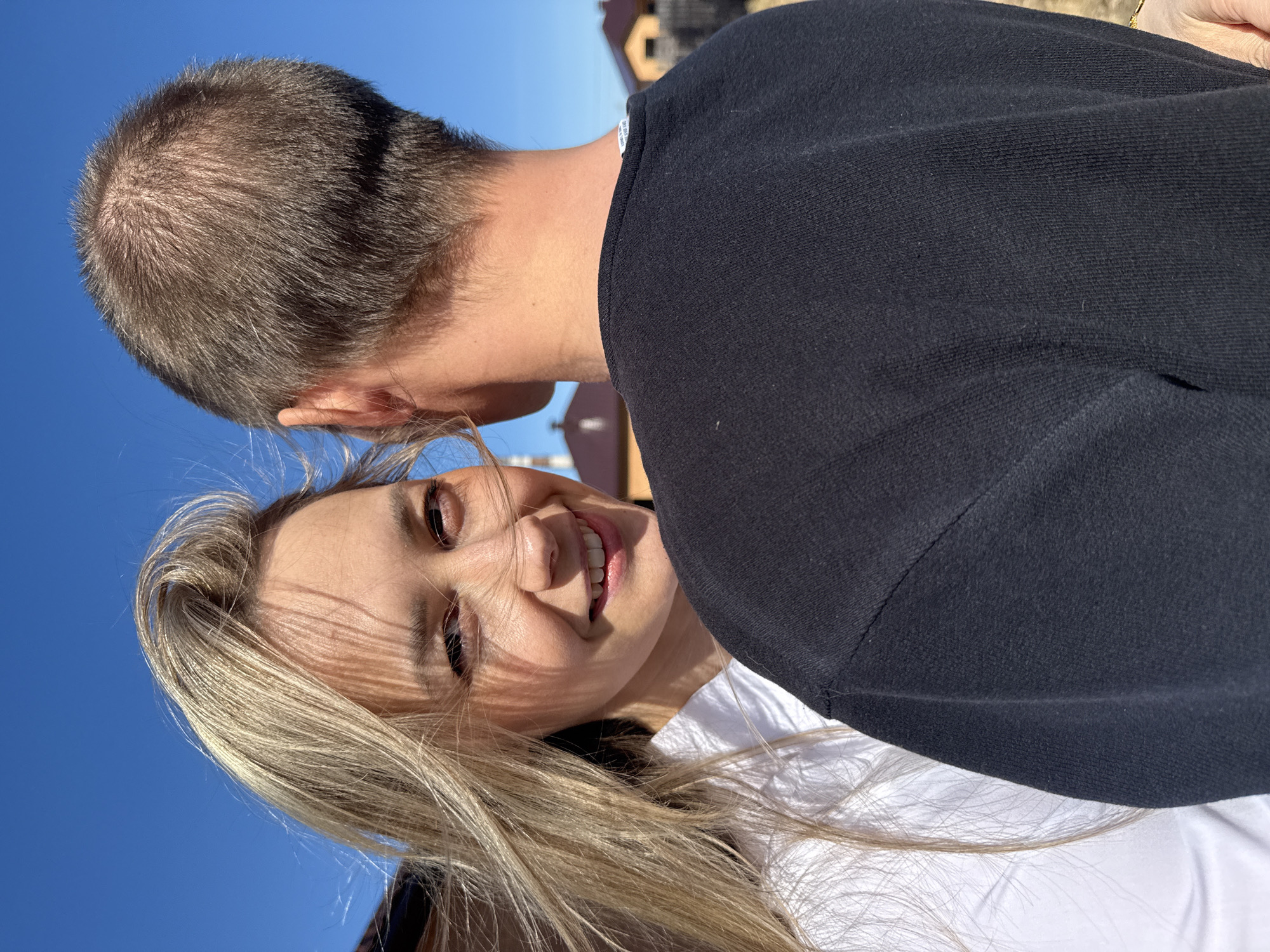 The image shows a woman with blonde hair and a white shirt, smiling at the camera. She is in close proximity to a man who is wearing a black sweater. The background is a clear blue sky with a building visible in the distance. The location is Chaikovsky, Russia.