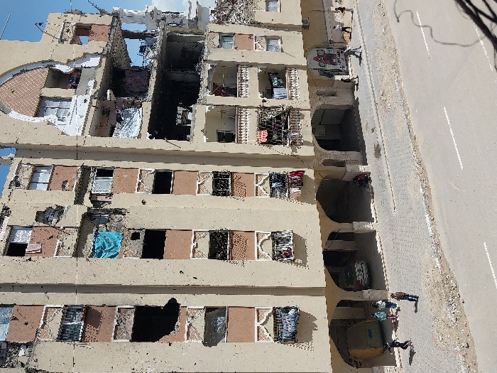 This image depicts a heavily damaged multi-story residential building in Khan Yunis, Palestinian Territory, under clear daytime conditions. The building's beige facade exhibits widespread small impact marks and significant structural damage, particularly on the upper right side where large sections of the exterior and interior walls are destroyed, exposing several apartments. Numerous windows are broken or missing, with some showing hanging personal items like clothing and curtains. A blue tarp hangs from a window on the left side. The ground floor features a series of arched openings, with one revealing a parked white car and another a figure seated near its entrance on the far right.
A grey paved sidewalk runs along the front of the building, separated from an asphalt road by a strip of dirt and debris. Approximately ten individuals are present on the sidewalk. On the left, a group of five people, including adults and possibly children, are gathered, with one person in a black shirt in motion and another in a brown jacket standing facing the street. Further right along the sidewalk, several other individuals are walking. A large, yellow-green cylindrical object rests near the group on the left. Overhead wires are visible in the bottom right corner, suggesting an elevated vantage point for the photograph.