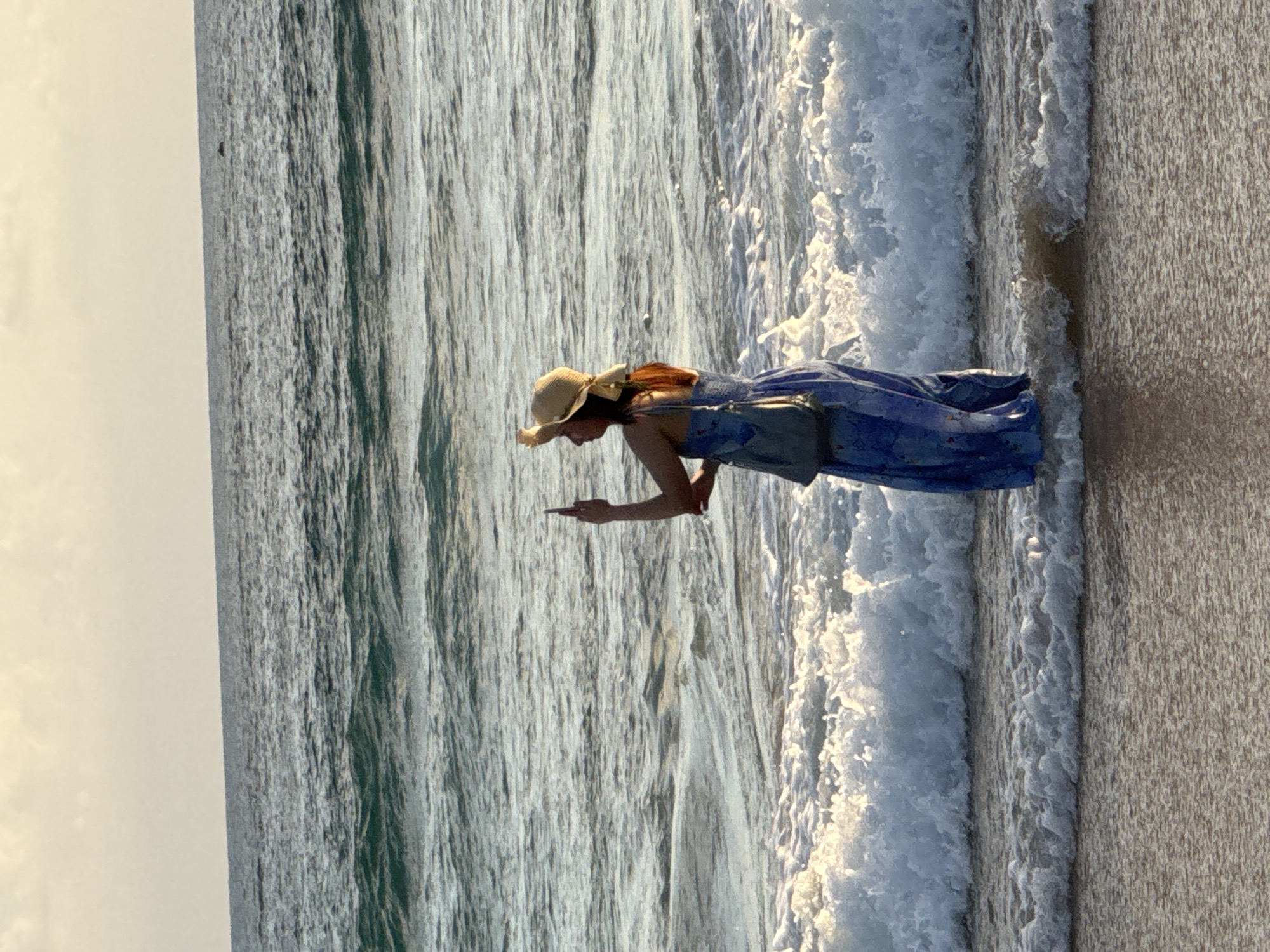 A person wearing a blue dress and a hat is standing in the ocean at Karon Beach in Thailand. The person is holding a phone and appears to be taking a picture or video of the ocean. Waves are crashing around the person. The sky is overcast.