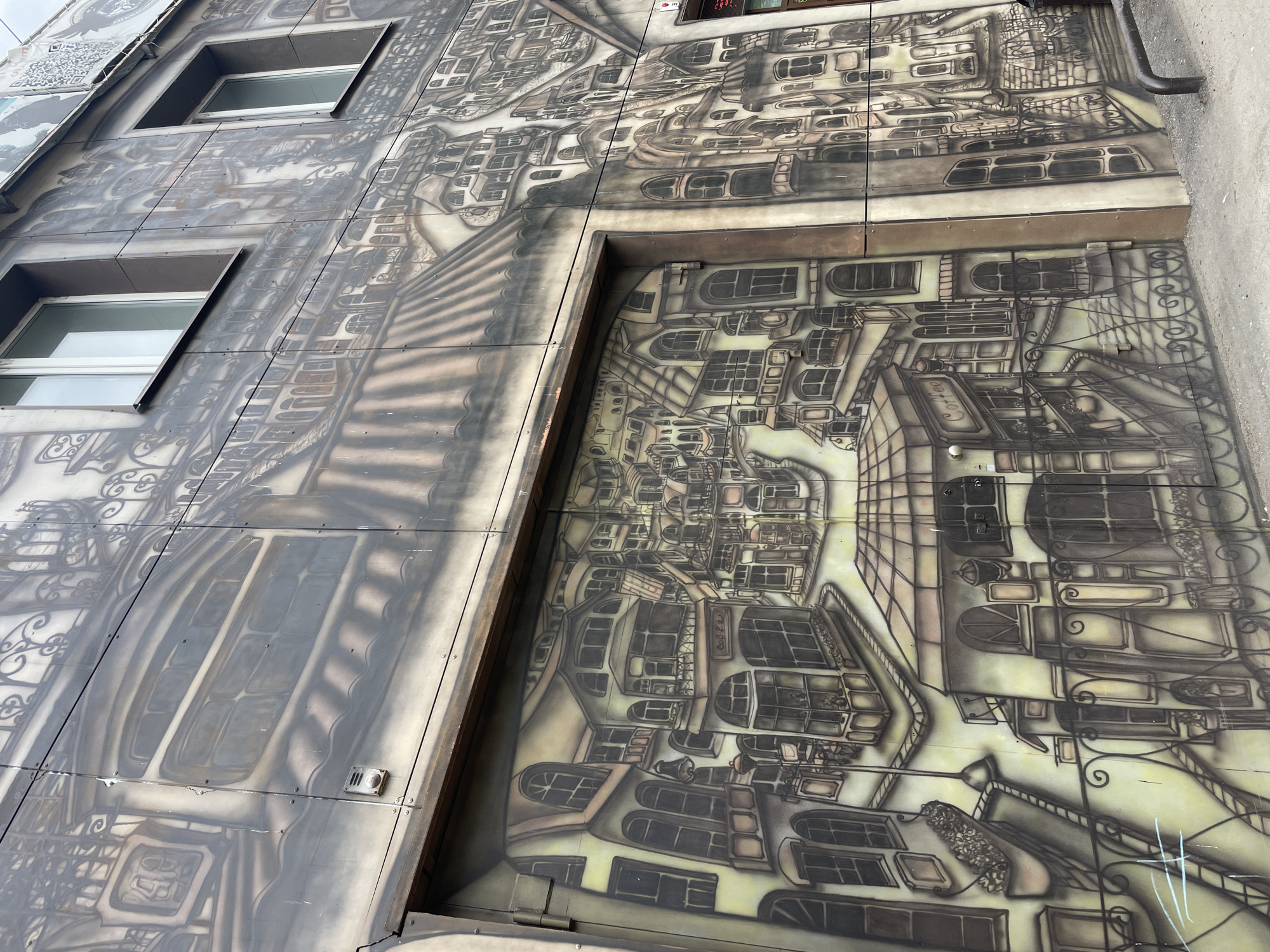Here's a description of the image:
The image shows a section of a building exterior in Krasnoyarsk, Russia. The facade is decorated with an elaborate, stylized mural in shades of gray and brown. The mural depicts an intricate cityscape featuring buildings with numerous windows, architectural details, and what appears to be wrought iron elements. The scene has a fantastical, almost surreal quality. The mural spans multiple panels of the building's cladding. Two rectangular windows with white frames are visible on the left side of the image. A utility pipe runs down the right side of the building. The image is taken at an angle, indicating a ground-level perspective. The overall aesthetic has a slightly aged or weathered appearance.