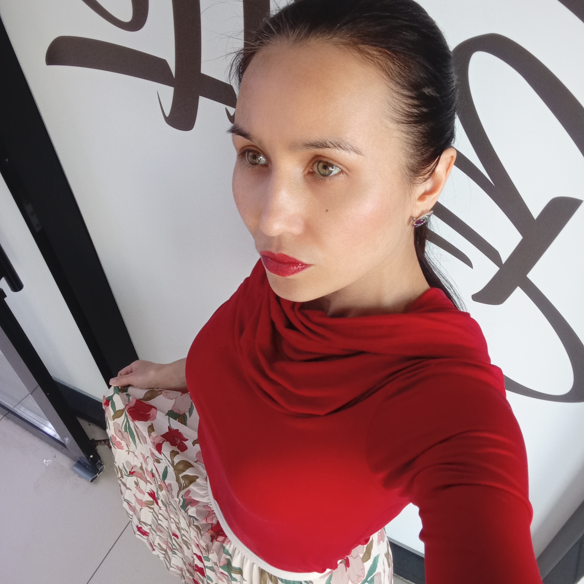 A woman stands indoors, appearing to pose for a photo. She wears a red top, a floral skirt, and has dark hair pulled back. She has red lipstick. Her gaze is directed off-camera to the left. Behind her is a white wall with dark brown abstract designs. A black doorframe is visible on the left. The location is suggested to be Izhevsk, Russia, but without further context, that can't be confirmed.