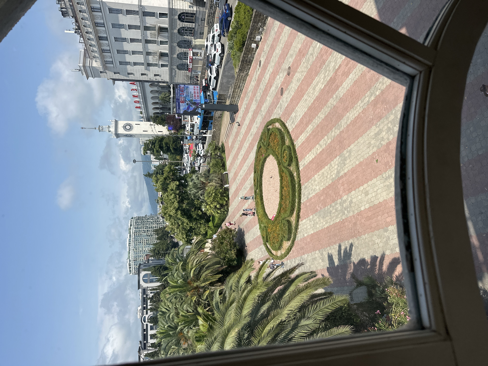 The image depicts a sunny day in the city of Sochi, Russia, as viewed from an interior window frame. In the foreground, a patterned pavement of red and white interlocking bricks stretches across a public square. A circular garden bed, filled with low green bushes and small yellow and orange flowers, is situated in the center of the square. Several palm trees, with their fronds casting shadows on the pavement, line the left side of the square. A few people are visible walking across the square.
In the background, a white building with a prominent clock tower stands tall, displaying the Cyrillic word "ВОКЗАЛ" (Vokzal, meaning "station") in large red letters above its entrance. Adjacent to the station is another large, white multi-story building with numerous windows. A busy street with cars and a blue bus is visible in front of these buildings. Beyond the structures, green hills and mountains can be seen under a partly cloudy sky. The scene appears to be a transportation hub or a central public area in Sochi.