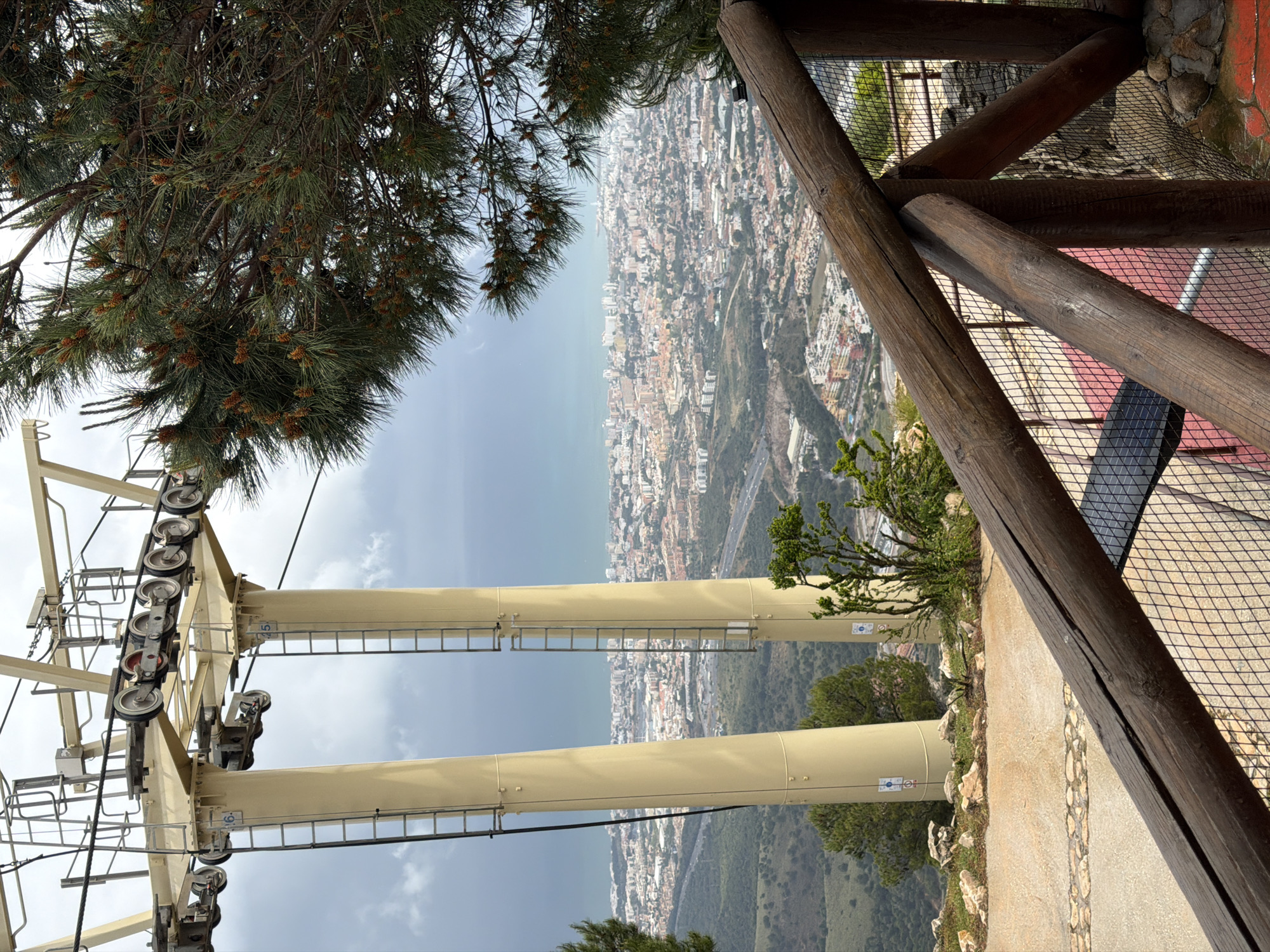The image depicts a view from a high vantage point in Arroyo de la Miel-Benalmádena Costa, Spain. The perspective is from an elevated structure, possibly a cable car station or lookout point. To the left, a portion of a cable car tower is visible, with wheels and support structures. In the foreground, a wooden railing borders the edge of the viewpoint.
Below, a city landscape stretches out toward the sea. Buildings and streets are discernible, and a highway winds through the terrain. The sky is overcast, suggesting a cloudy day. Vegetation is present in the foreground and on the slopes beneath the city, indicating a hilly or mountainous terrain.