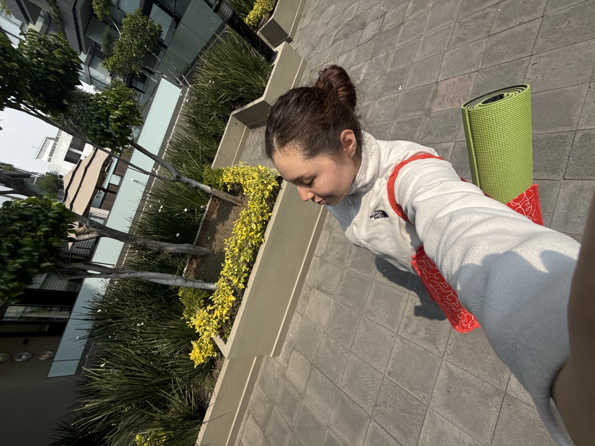 A high-angle medium shot depicts a young woman standing on a tiled walkway in Mexico City, Mexico. She is wearing a white fleece jacket with a red shoulder bag and carrying a rolled-up green yoga mat. Her hair is pulled back in a bun. Along the left side of the image, there is a low concrete planter containing various plants, including tall grasses and yellow flowering shrubs. Behind the planter, a multi-story building can be seen. The day appears to be sunny and bright.