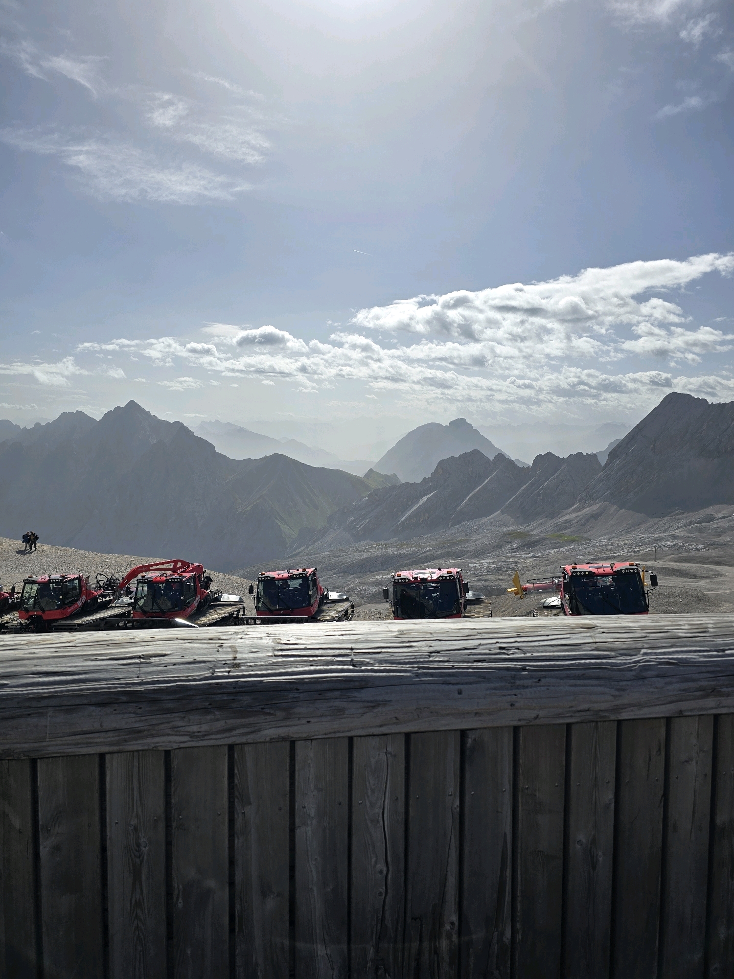 The image captures a scenic mountain landscape with a clear, bright sky and scattered clouds. In the foreground, a rustic wooden fence runs horizontally across the bottom of the frame. Behind the fence, a line of several red snow grooming vehicles is parked on a gravelly surface. The vehicles are positioned facing forward, with their tracks visible. In the mid-ground, a vast expanse of rugged mountains stretches into the distance, appearing hazy due to atmospheric perspective. Two small figures are visible walking on the slope between the snow groomers and the mountains. The scene suggests a location in the Bavarian Alps, likely near the Zugspitze, Germany's highest peak, which is accessible from Garmisch-Partenkirchen. The overall impression is one of a clear, sunny day in a high-altitude mountainous region, potentially a ski resort area during the summer months or off-season.