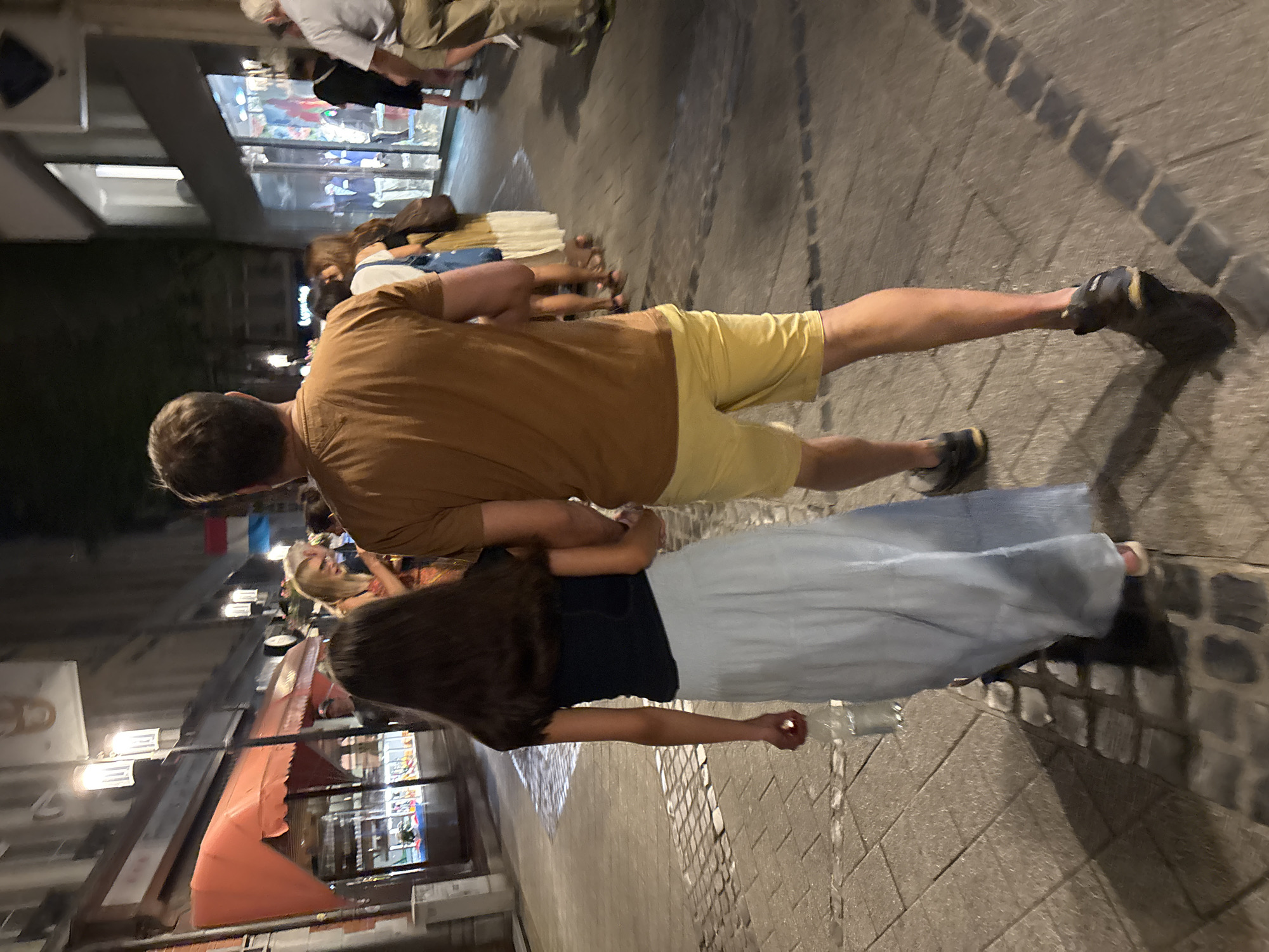 The image captures a view from behind two people walking on a cobblestone street at night in Luxembourg City. A man in a brown t-shirt and yellow shorts walks ahead, holding hands with a young girl in a dark blue top and a light blue skirt. The girl is carrying a clear plastic bottle with a red cap in her left hand. They are walking on a street lined with buildings, one of which has an orange awning with lights. Other people are visible in the background, walking in the same direction. The street lighting casts shadows on the cobblestones. The overall impression is a casual evening stroll through a European city.