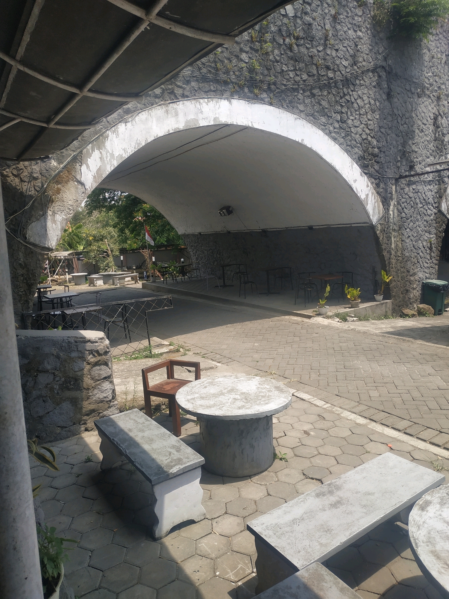 The image shows an outdoor seating area under a large arched structure, possibly a cafe or restaurant, located in Patrang, Indonesia. The foreground features several concrete benches and a circular table with a wooden chair. The ground is paved with interlocking hexagonal tiles. Beyond the immediate seating area, under the arch, are more tables and chairs, with a stone wall on the right side and trees and foliage visible in the background. A green trash bin is also present on the right. The overall scene suggests a relaxed outdoor environment, likely designed for patrons to enjoy food and drinks.
