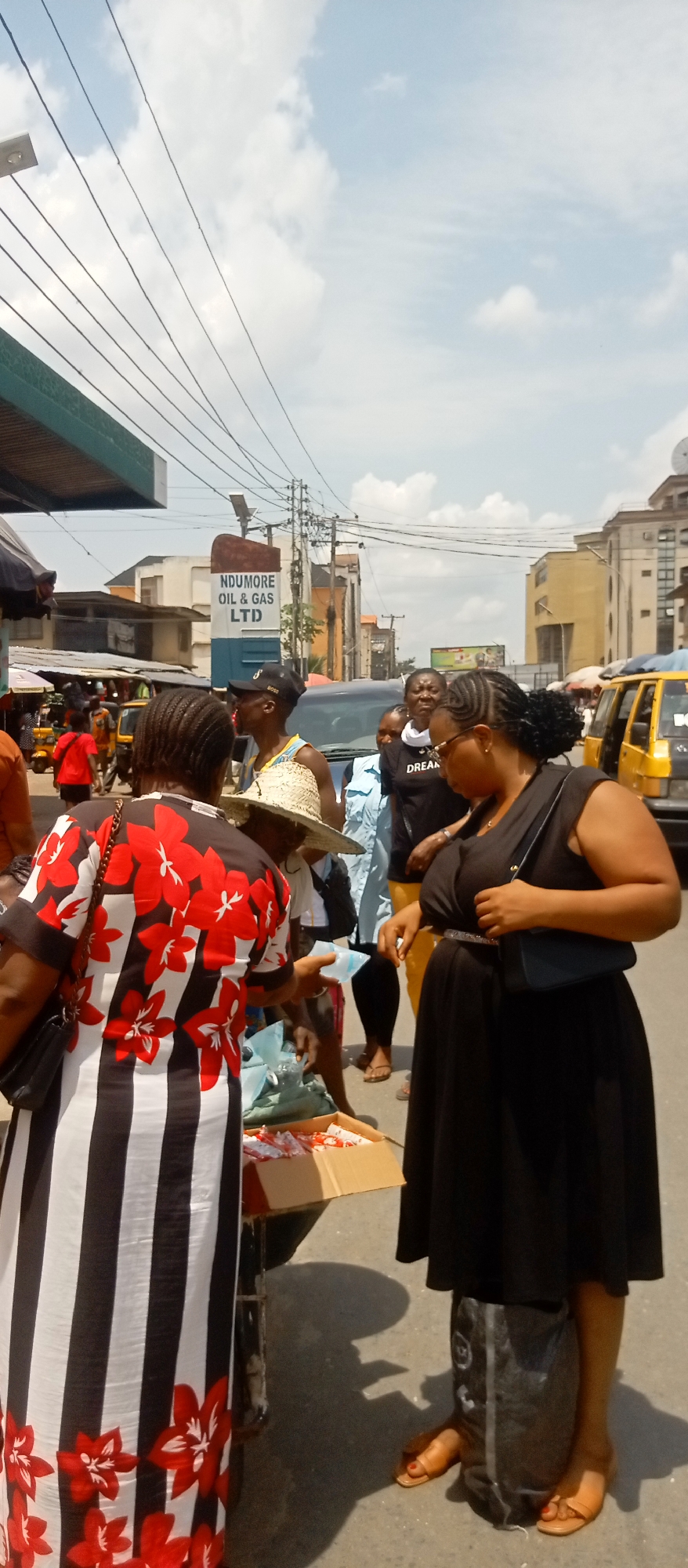 This media file depicts a busy outdoor street scene in Onitsha, Nigeria, captured on a bright, cloudy day, likely midday or early afternoon. The setting appears to be a market or commercial area, bustling with people and activity.
In the foreground, two women are central. The woman on the left, with her back to the camera, wears a distinctive white dress adorned with black vertical stripes and red floral patterns, carrying a black shoulder bag. She is leaning towards a low table or stand, seemingly engaged in a transaction involving small packaged items displayed in a cardboard box. To her right, another woman in a black sleeveless dress and dark belt, carrying a black shoulder bag, stands in profile, looking down. Her feet are visible, wearing light brown sandals, and she is next to a black bag on the ground.
Several other individuals are visible in the mid-ground, including a man wearing a straw hat and a black cap (with faint "BOSS" text) and another person in a black t-shirt with the word "DREAM" printed on it. Vehicles, including typical yellow taxi cabs and a dark SUV, are present on the road, indicating active traffic.
The background reveals a street lined with various buildings and utility poles with numerous power lines crisscrossing the sky. A prominent sign for "NDUMORE OIL & GAS LTD" is visible. The scene portrays a vibrant urban environment characteristic of a busy Nigerian street or market.