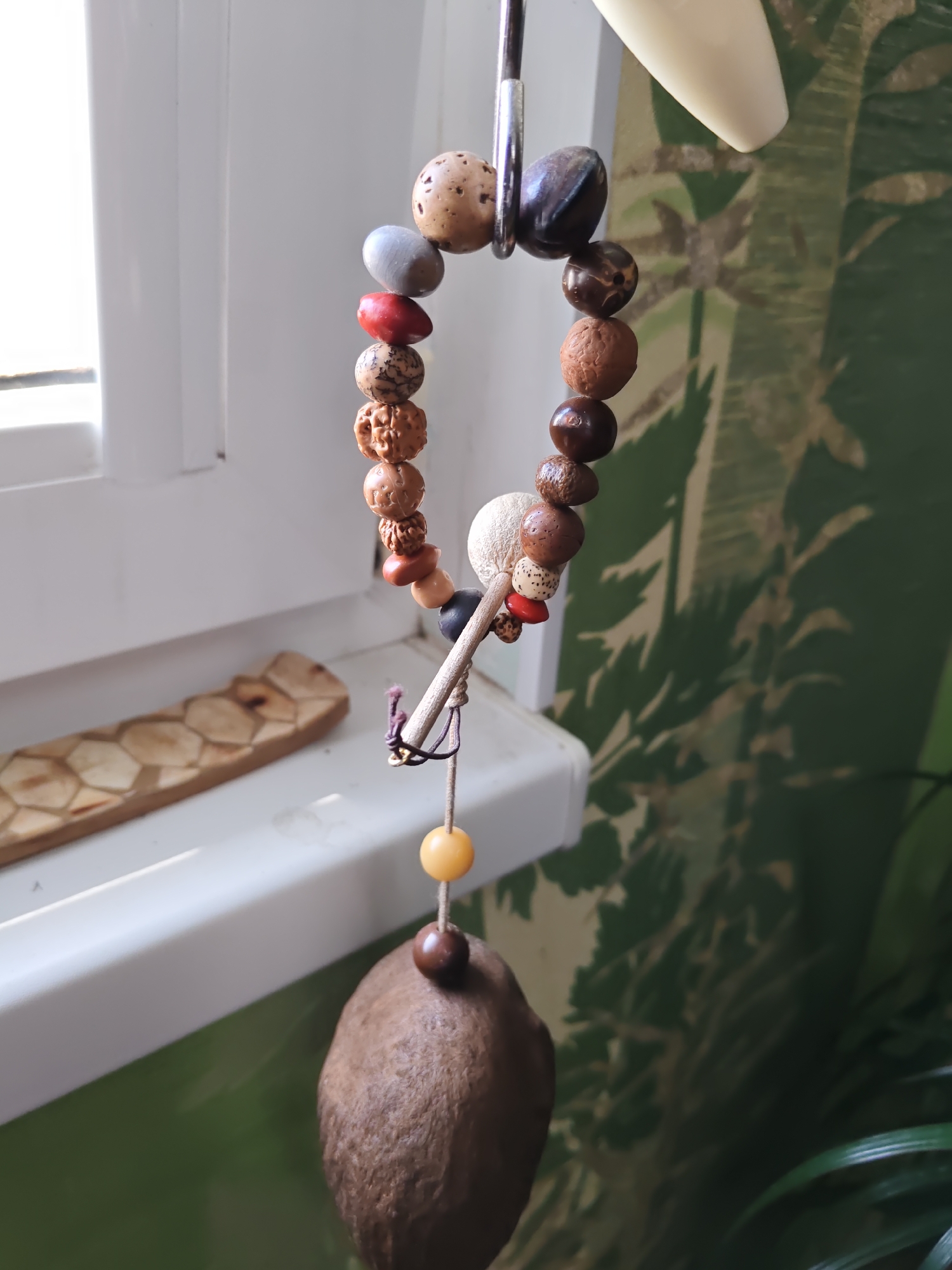 A beaded ornament with various natural materials hangs from a metal hook near a window. The ornament features a collection of beads made from seeds, stones, and possibly carved wood, in shades of brown, beige, red, and grey. A larger, textured brown object hangs at the bottom. To the left, on the windowsill, is a carved wooden object with a hexagonal pattern. The background is a green wall with a pattern of leaves and possibly palm trees. The scene is lit by natural light coming from the window. There are no people present in the image.
