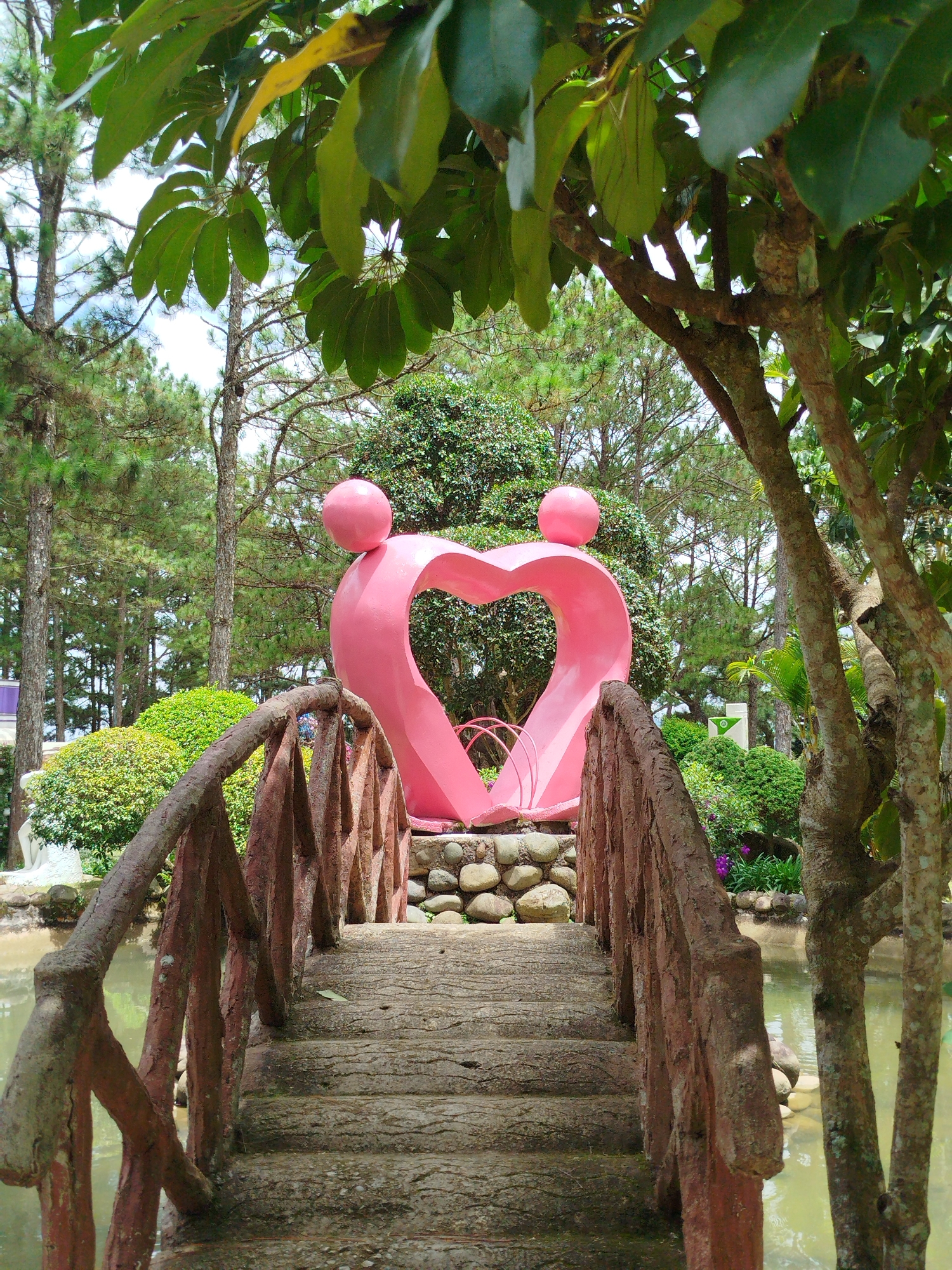 The image depicts a charming park scene, likely within the Lâm Viên Highlands of Đà Lạt, Vietnam. In the foreground, a rustic, red-brown stone bridge with weathered railings arches over a calm body of water. The bridge's walkway is constructed of textured stone slabs. Beyond the bridge, a vibrant pink sculpture, resembling two figures forming a heart shape with rounded heads, stands as the central focal point. The sculpture is situated on a bed of river stones. Lush greenery surrounds the sculpture, including manicured bushes and tall pine trees characteristic of the region. The water in the pond reflects the surrounding foliage and the overcast sky. The overall atmosphere is tranquil and picturesque, suggesting a public garden or a romantic spot within a larger resort or attraction. There are no people visible in the frame.