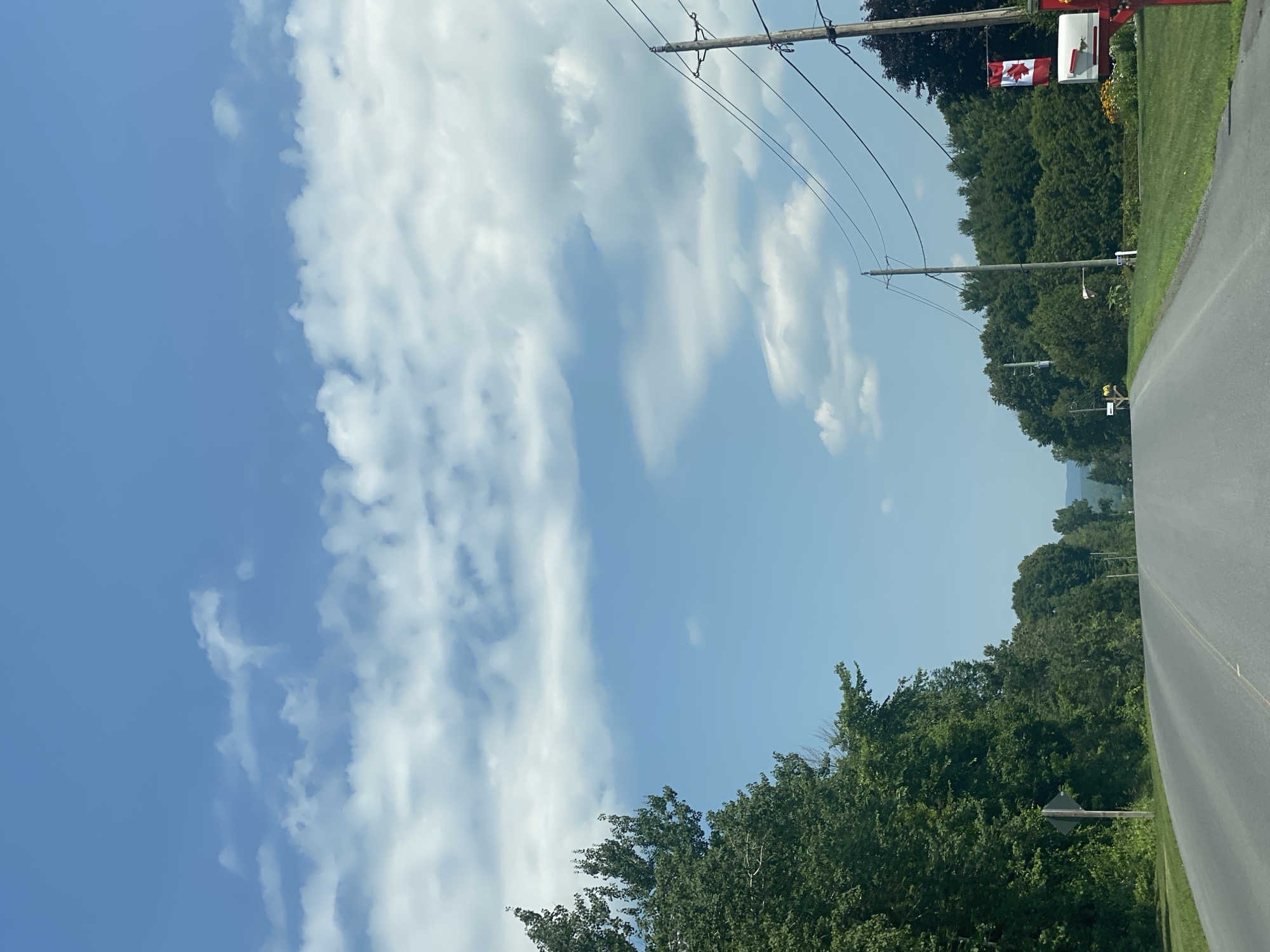The image shows a straight asphalt road passing through a rural area with trees lining both sides. The sky is a clear blue with scattered white clouds. Power lines run along the right side of the road, supported by wooden poles. A Canadian flag is attached to one of the poles, near a red mailbox. The area appears to be residential, with manicured lawns and gardens visible. The overall scene depicts a serene summer day in a Canadian countryside setting.