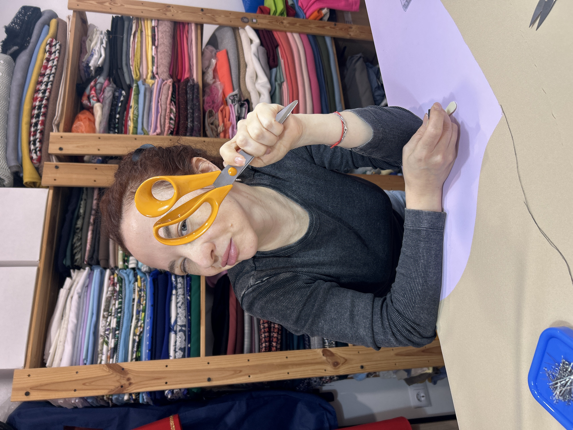 A woman with reddish-brown hair is looking at the camera while holding large yellow-handled scissors up to her eye, peering through the handle opening. She is wearing a gray long-sleeved shirt with a slightly textured pattern. Her right hand is holding a piece of tailor's chalk against a light purple piece of material laid over a beige surface. The background is filled with shelves stacked with various fabrics, suggesting a sewing room or studio. A blue container with sewing pins sits on the beige surface to the right. The location is an indoor space, potentially a home or studio, in an unknown city.