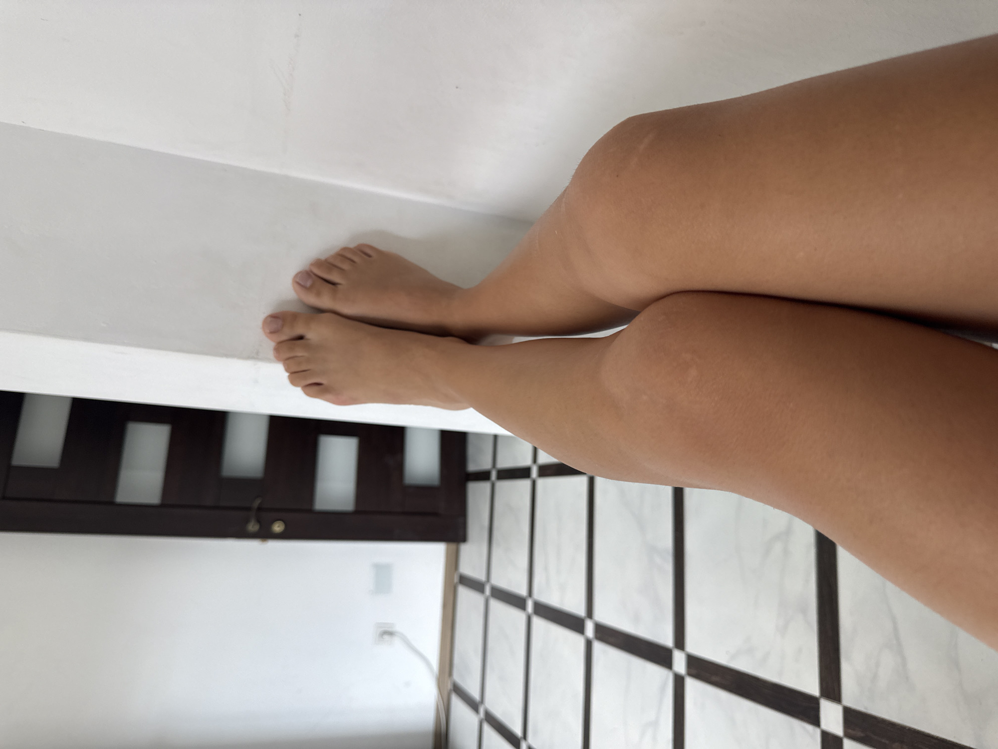 A first-person perspective shot shows a pair of bare legs resting against a white wall. The legs are tanned and show some faint white marks, possibly from scars or stretch marks. The feet are visible at the top of the frame, with toes pointed upwards. The floor in the background is tiled with a black and white checkered pattern. To the left of the legs, a dark brown door with frosted glass panels is visible. A white electrical outlet with a cord is also on the wall to the left. The overall scene appears to be indoors, likely in a residential setting within the city of Chișinău, Moldova. The lighting is natural and bright, indicating daytime. No other individuals are present in the frame.