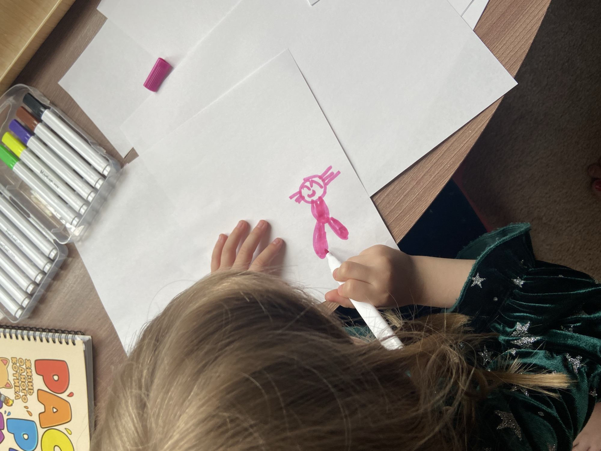 A child is drawing a pink stick figure on a white sheet of paper. The child is wearing a green velvet dress with silver stars. On the table are a coloring book, a set of markers in a clear plastic case, a pink marker cap, and several sheets of white paper. The image appears to be taken indoors. The location is unknown.