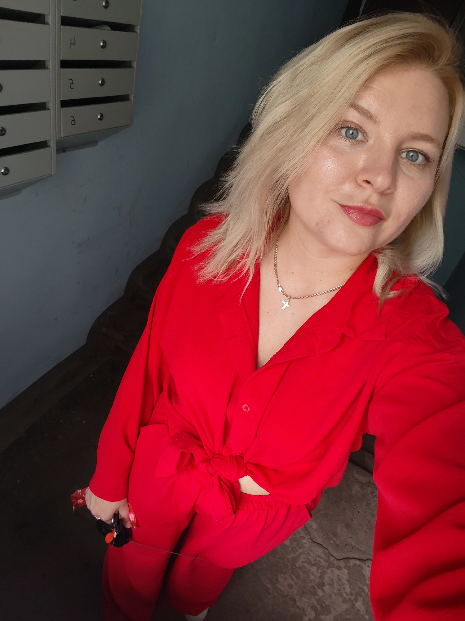 A blonde woman with blue eyes and light makeup is taking a selfie in a stairwell. She is wearing a red collared shirt, tied at the waist, and matching red pants. A thin gold chain with a cross pendant is visible around her neck. She is holding a small, black object with an orange accent in her right hand. Behind her, on the left wall, are several light gray mailboxes, some with letters written on them. The stairwell appears to be dimly lit, with wooden stairs descending in the background. The overall impression is that of an indoor residential building, possibly an apartment complex, in Izhevsk, Russia.