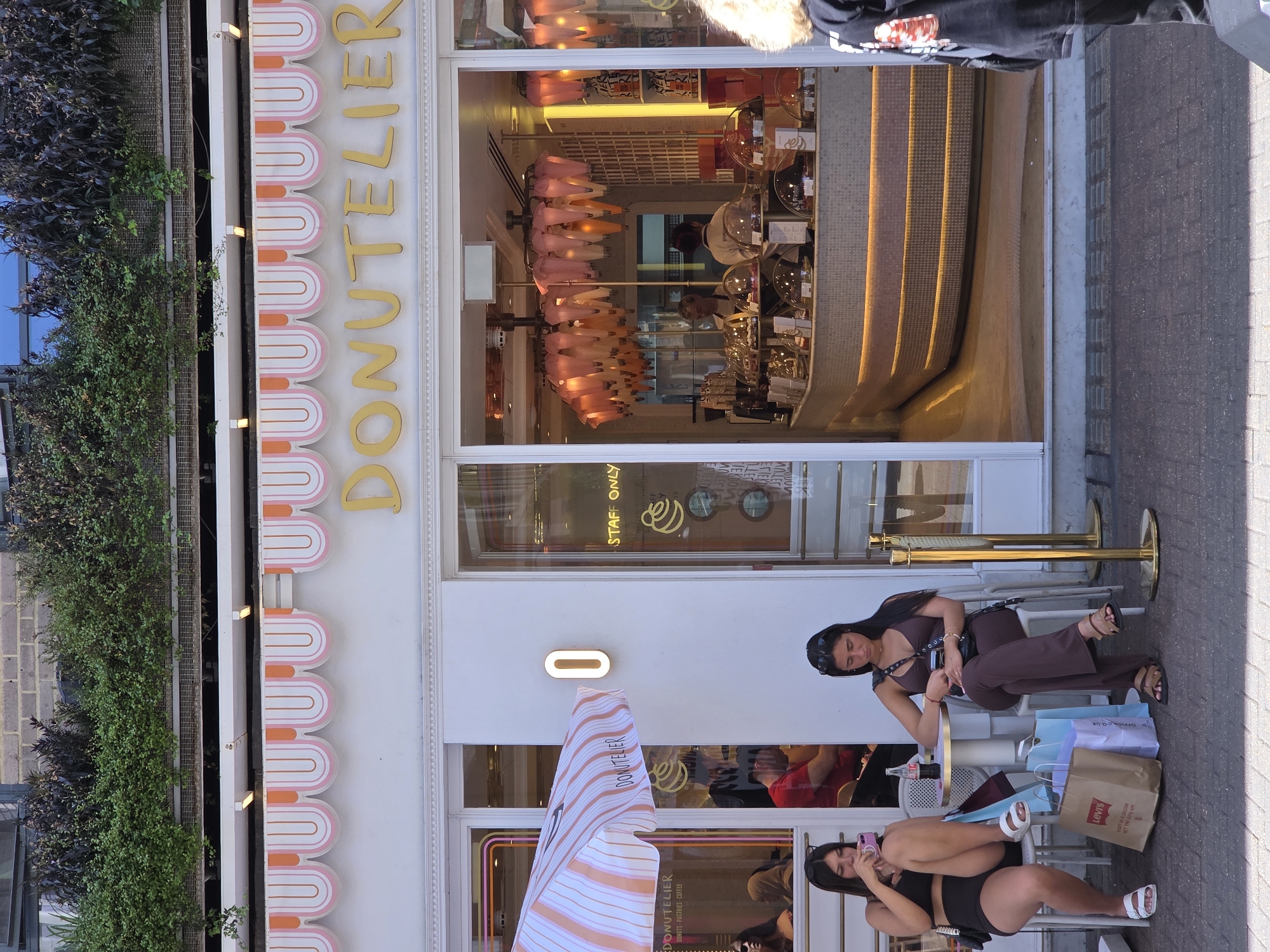 Here is a detailed description of the image:
**Setting:** The image shows the exterior of a store named "Donutelier" in London, United Kingdom. The store's facade is white with an orange and white scalloped awning. The sign reads "DONUTELIER" vertically in gold letters, and a smaller sign indicates "DONUTS, PASTRIES, COFFEE". There's a striped umbrella also with the "Donutelier" name. The shop windows reveal the interior with a modern design, including pink hanging fixtures and a curved counter with golden accents. A sign on the window reads "Staff Only". Plants are growing up the adjacent building.
**People:** Two young women are sitting at a small table outside the store. One woman is wearing brown clothing and appears to be looking at her phone. The other woman, wearing black, is also looking at her phone and sitting in a relaxed posture. Shopping bags, including one from Levi's and one from Office.co.uk, are placed near their feet. A third person is partially visible walking by the store.
**Objects:**
* "Donutelier" Sign and awning
* Tables and chairs
* Shopping bags
* Smartphones
* Storefront window display with interior decor
**Actions:**
* People sitting and using their phones
* People walking by
**Overall Impression:** The scene captures a moment of everyday life in London, with people relaxing outside a cafe or bakery. The bright, modern design of "Donutelier" stands out, and the image suggests a trendy, upscale establishment.