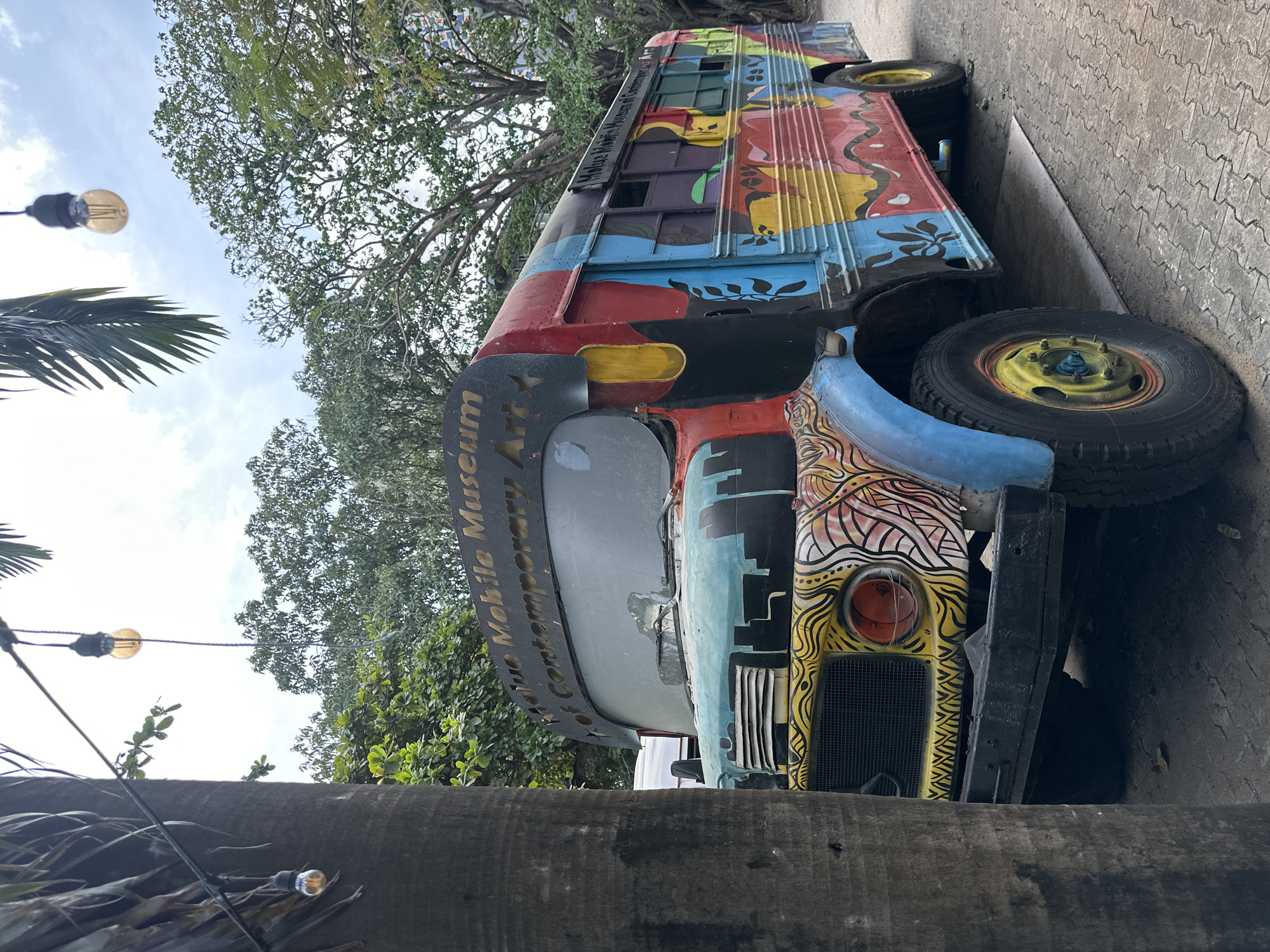 A bus parked on a concrete street in Lagos, Nigeria. The bus is painted with colorful graffiti. The words "Mobile Museum of Contemporary Art" are painted on the front of the bus. Trees and string lights are visible in the background.
