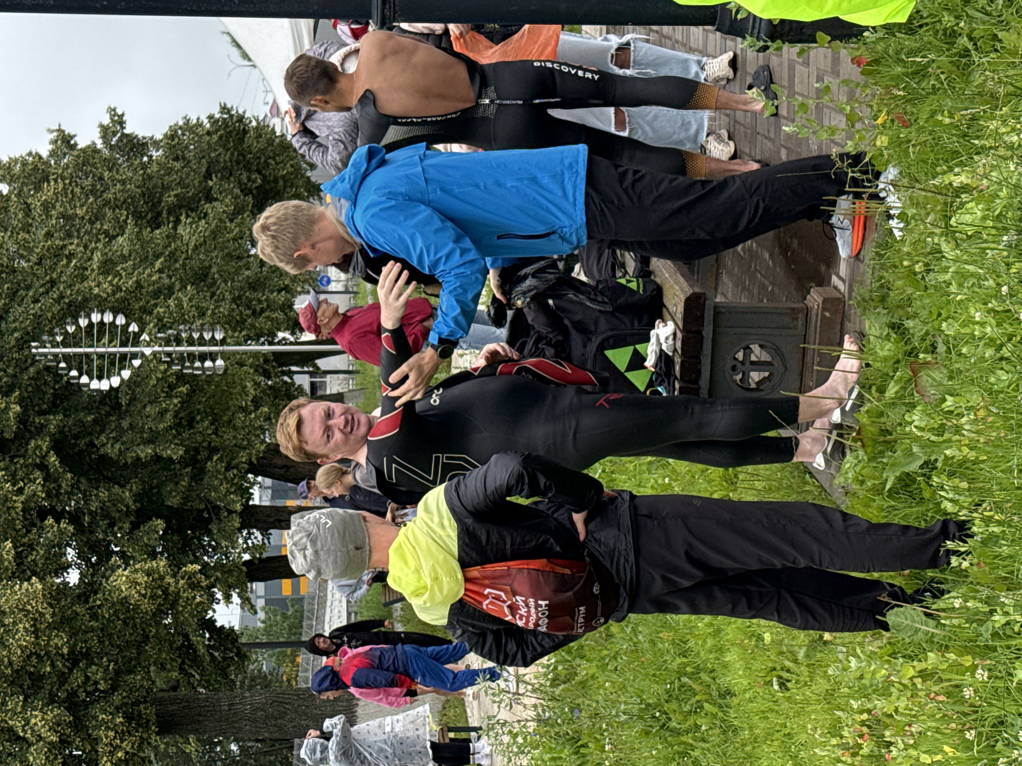 A group of people, some wearing wetsuits, are gathered outdoors in Votkinsk, Russia, likely near a body of water for a sporting event. The foreground features a man wearing a grey beanie and a neon yellow and black jacket with a red drawstring bag. He is standing next to a man in a black wetsuit with red accents, who is partially unzipping the suit. To the right, another man is wearing a blue jacket and black pants, his back to the camera. In the background, several other individuals are visible, some in rain gear. The environment includes lush green grass, trees, and a paved path. A metal bench is positioned in the middle ground. The overall atmosphere suggests a pre or post-event gathering for a triathlon or similar water sport.