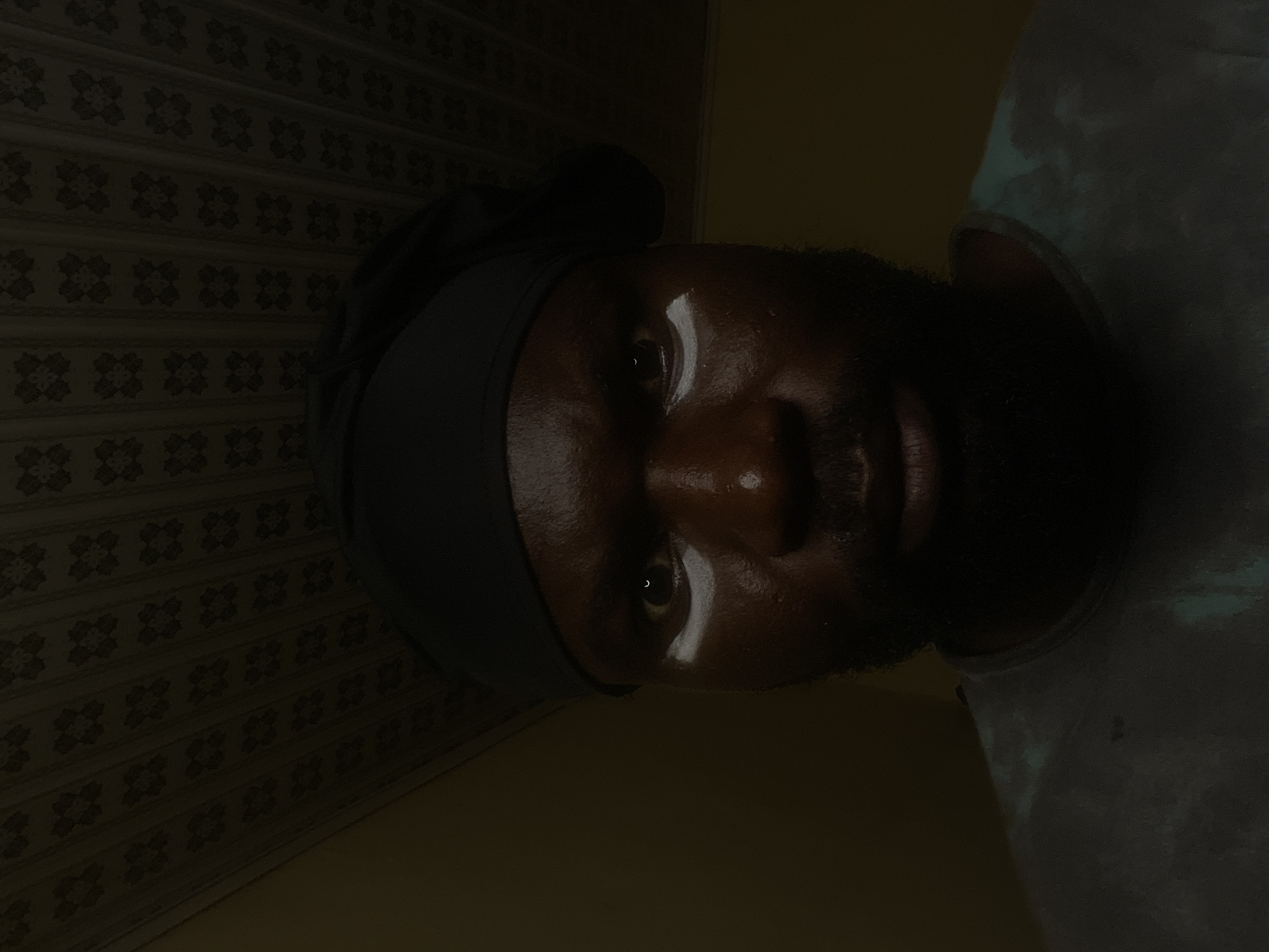 A dark-skinned person wearing a dark-colored head covering and a light-colored shirt is viewed at close range. Their eyes are open, and they are looking directly at the camera. The lighting is dim, casting shadows on their face. In the background, there is a patterned wallpaper. It is not possible to determine if this photograph was taken in Gura Top, Nigeria.