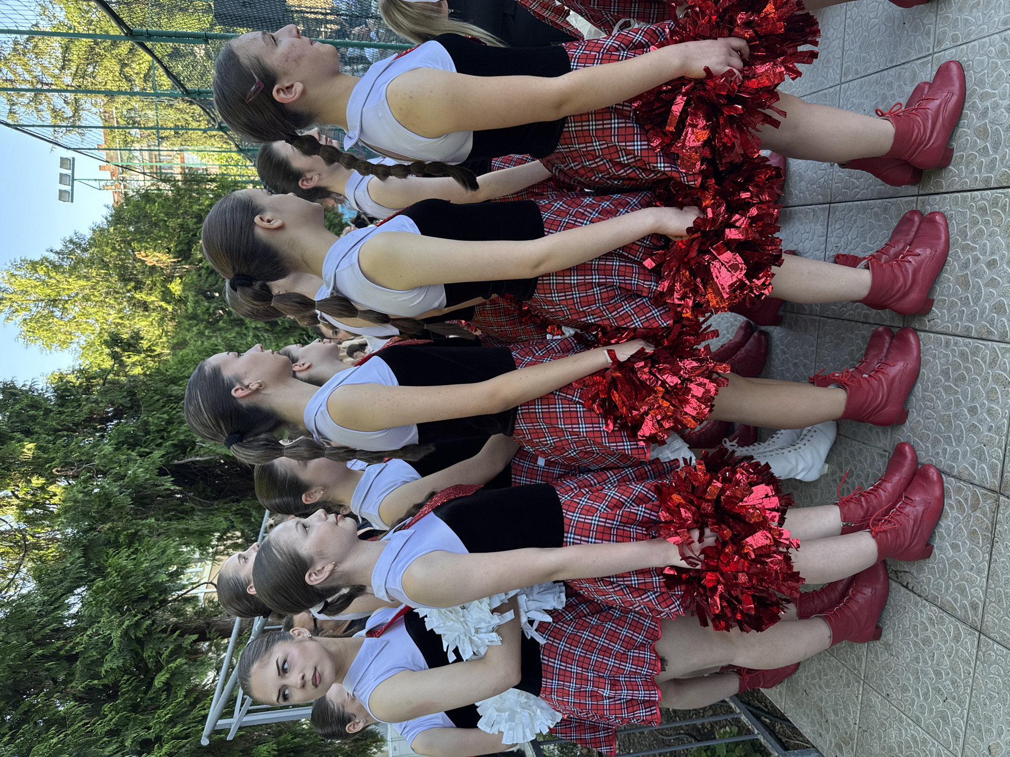 A group of young women, dressed in matching cheerleading uniforms consisting of a black sleeveless top with a white collar and red tie, a red and black plaid skirt, and red lace-up boots, stand in formation on a tiled outdoor area. They are holding white and red pom-poms. The background features trees, a chain-link fence, and a building. This scene is located on the grounds of the Pleven Regional Library in Pleven, Bulgaria.