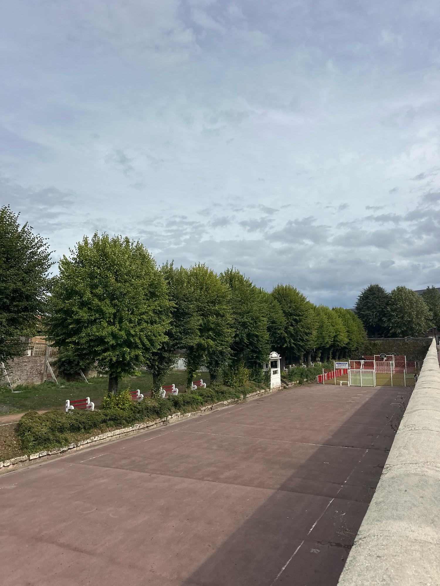 The image captures a view of the pétanque court in the city of Roye, France, on a cloudy day. In the foreground, a large, reddish-brown court occupies the majority of the frame, with a light gray, textured wall on the right. To the left of the court, a line of trees with lush green foliage runs along a grassy area. Several white benches with red seats are placed along the edge of the grassy area. In the background, there is a colorful sports court with red and green fencing and a white structure that appears to be a scoreboard or changing room. The sky above is filled with a mix of white and gray clouds.