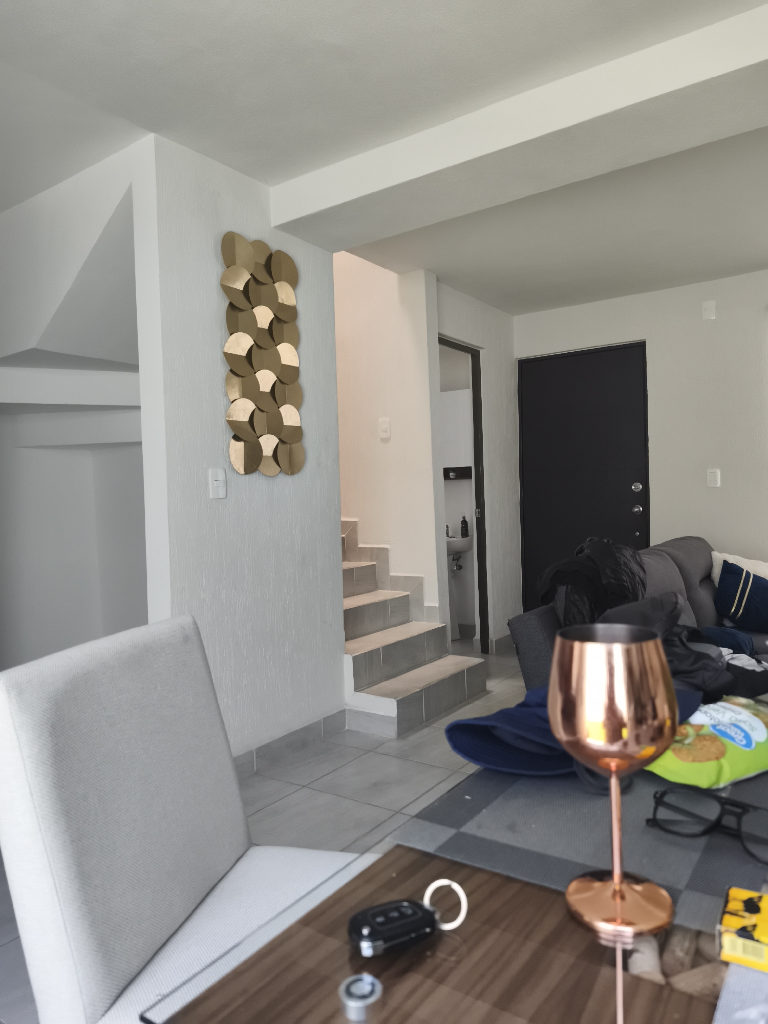 The interior of a residential space in El Carmen, Mexico, is shown. The room features tile flooring and white walls. A staircase with gray steps leads to an upper level. The wall next to the stairs is decorated with a vertical art piece composed of gold and brown discs. A doorway leads to a small washroom with a sink. A dark-colored door is also visible, possibly an exterior door.
In the foreground, a table holds a car key, glasses, a package of cookies, and a rose-gold colored wine glass. A light-gray chair is positioned next to the table. In the background, a gray sofa is visible with pillows. There are no people present in the frame.