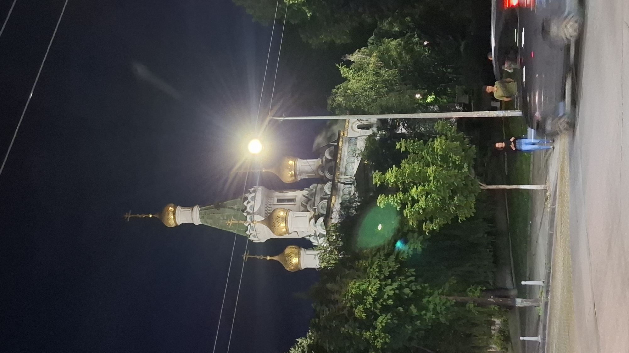 At night in Sofia, Bulgaria, a brightly lit street lamp illuminates the facade of the Saint Nicholas the Miracleaker Russian Church. The church, an architectural masterpiece with golden domes and intricate white detailing, is partially obscured by lush green trees. In the foreground, a dark-colored SUV drives by, its motion blurred by the low light. Two individuals stand on the sidewalk near the church, observing the scene.