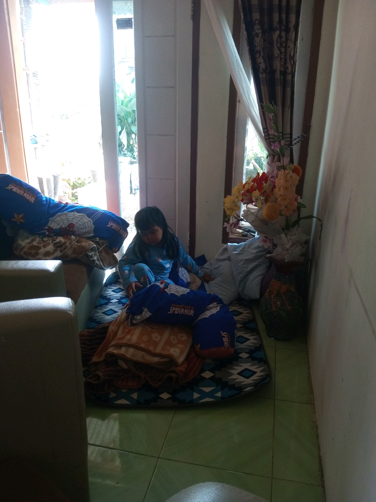 A young girl wearing a blue Spider-Man pajama set sits on a rug on the floor in a room that appears to be a living room or play area in Krasak, Indonesia. She is looking down, possibly playing with something or resting. The rug is patterned with blue and white diamonds. Next to her are several blue Spider-Man pillows and folded blankets. Behind her, a light-colored couch is visible. To the right of the girl, a window with a patterned curtain is open, allowing natural light to enter the room. In front of the window, a vase filled with artificial flowers sits on the floor. The floor is tiled in a green color. The overall impression is of a casual, indoor setting.