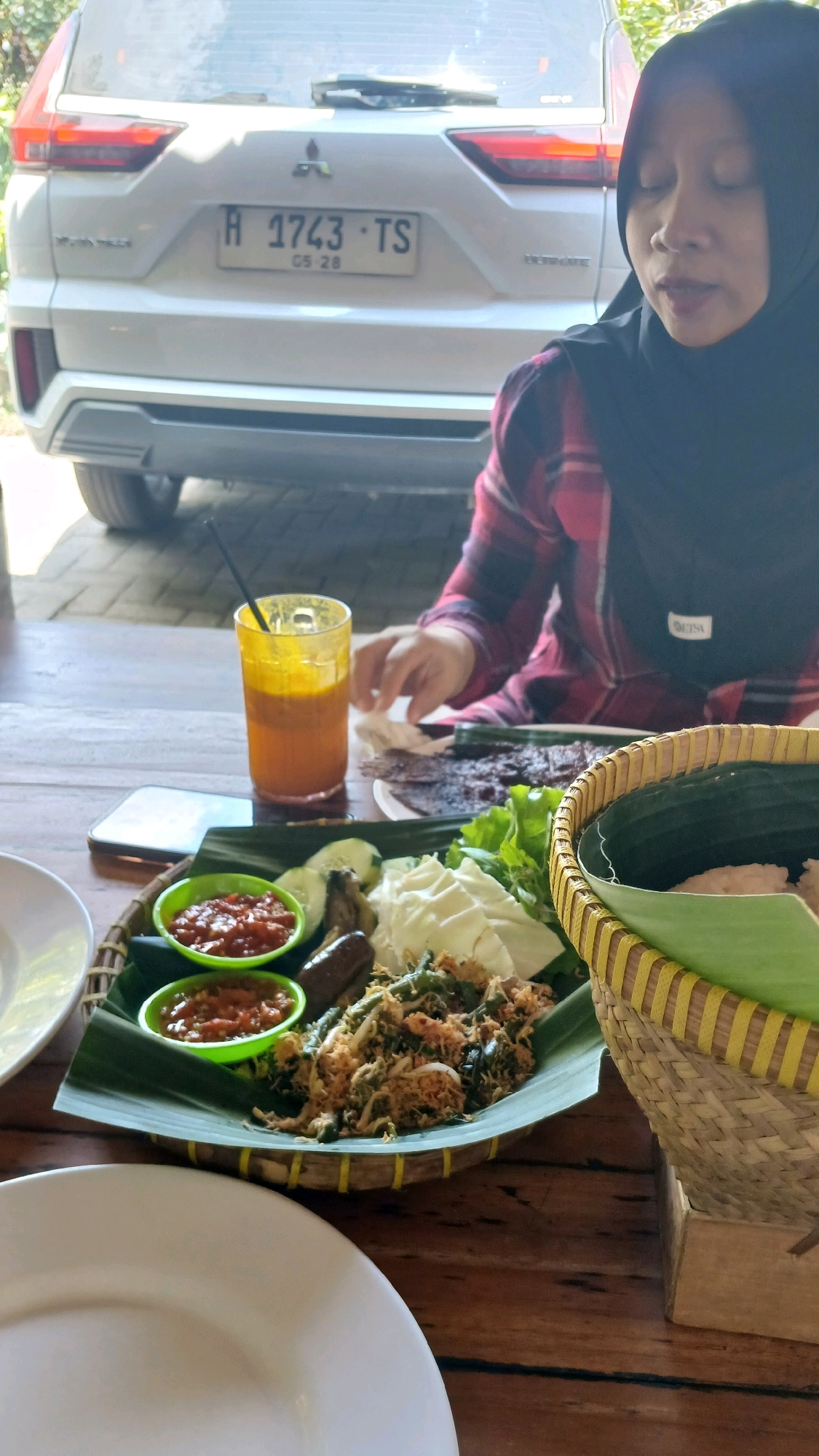 The image shows a woman wearing a black hijab and a red and black plaid shirt. She is seated at a wooden table outdoors, with a white Mitsubishi Xpander car visible in the background. The car's license plate reads "H 1743 TS". On the table in front of the woman is a meal served on a banana leaf within a woven basket. The meal consists of rice, vegetables, and two small bowls of red chili sauce. A glass of orange liquid with a straw is also on the table. The scene appears to be set in a restaurant or outdoor eating area in the city of Madiun, Indonesia, given the Indonesian license plate format and the food presentation style.