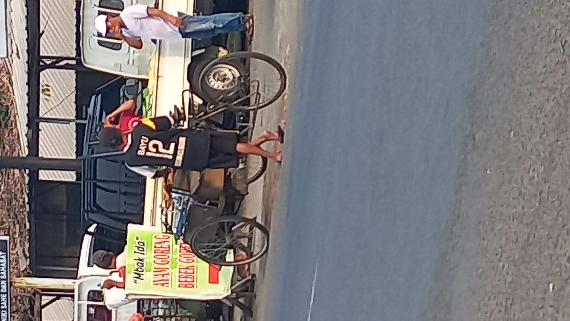 A medium shot captures a street scene in Mangunsari, Indonesia. In the foreground, a bicycle rickshaw, commonly known as a becak, is parked. A young man wearing a black and yellow jersey with the number "12" and the name "BAYU" on the back stands beside it, facing away from the camera. He is wearing black shorts and flip-flops. To the left of the becak, a green and white sign advertises "Mbak Ida" with "AYAM GORENG" (fried chicken) and "BEBEK GORENG" (fried duck) written on it.
Behind the becak, a black pickup truck is partially visible. To the right, a white truck is parked, and a man in a white t-shirt and blue jeans stands by it, looking to his left. Another person, wearing a red shirt, is partially visible behind the pickup truck, seemingly interacting with the man in the black and yellow jersey. The background suggests a street lined with various structures, possibly stalls or shops, under a covered area. The ground is paved, and the overall lighting indicates it is daytime.