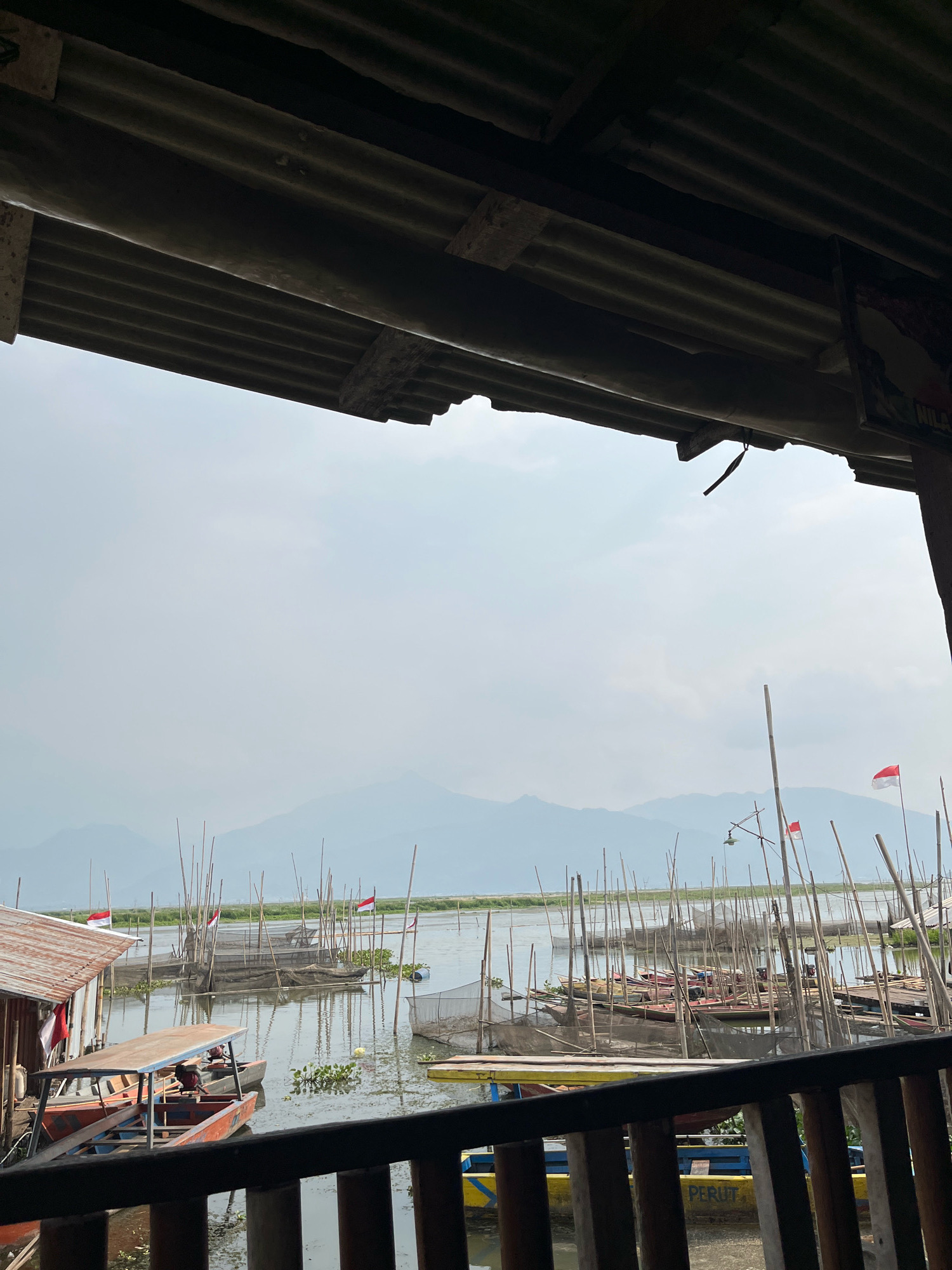 The image depicts a view from under a corrugated metal roof, looking out over a body of water in Tuntang, Indonesia. Numerous wooden poles emerge from the water, many of them supporting fishing nets or small structures. Several boats are moored or docked, some appearing to be fishing vessels, while others might be used for transport. Two Indonesian flags, red and white, are visible on poles scattered amongst the structures. In the distance, hazy mountains form the horizon under a cloudy sky. The foreground shows a wooden railing. The overall scene suggests a coastal or lakeside fishing community.