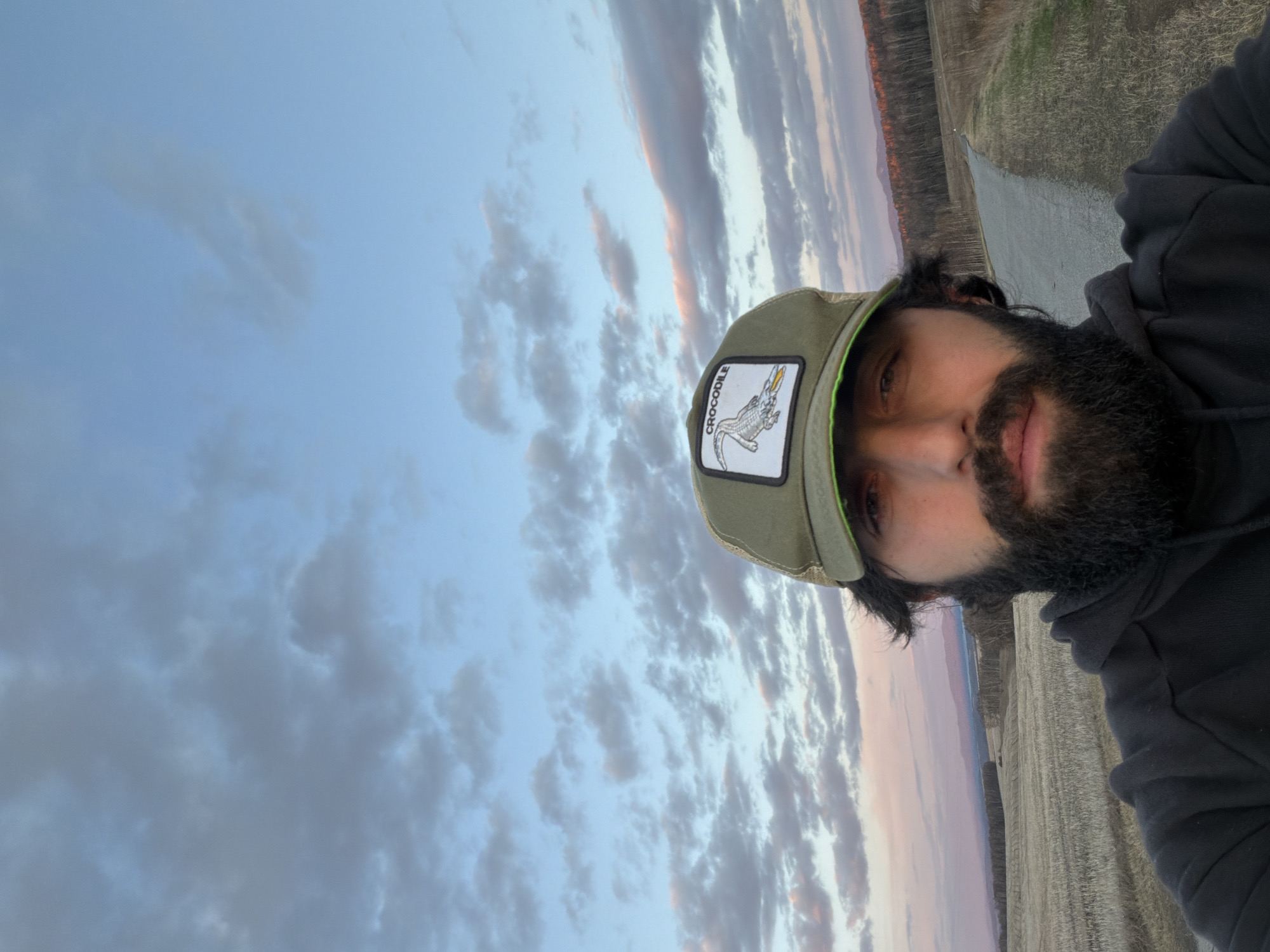 A person with a dark beard and wearing a "Crocodile" hat and a black hoodie is taking a selfie. The background features a sky with light clouds and a landscape with trees and a road. The location is Chesnoki, Russia.