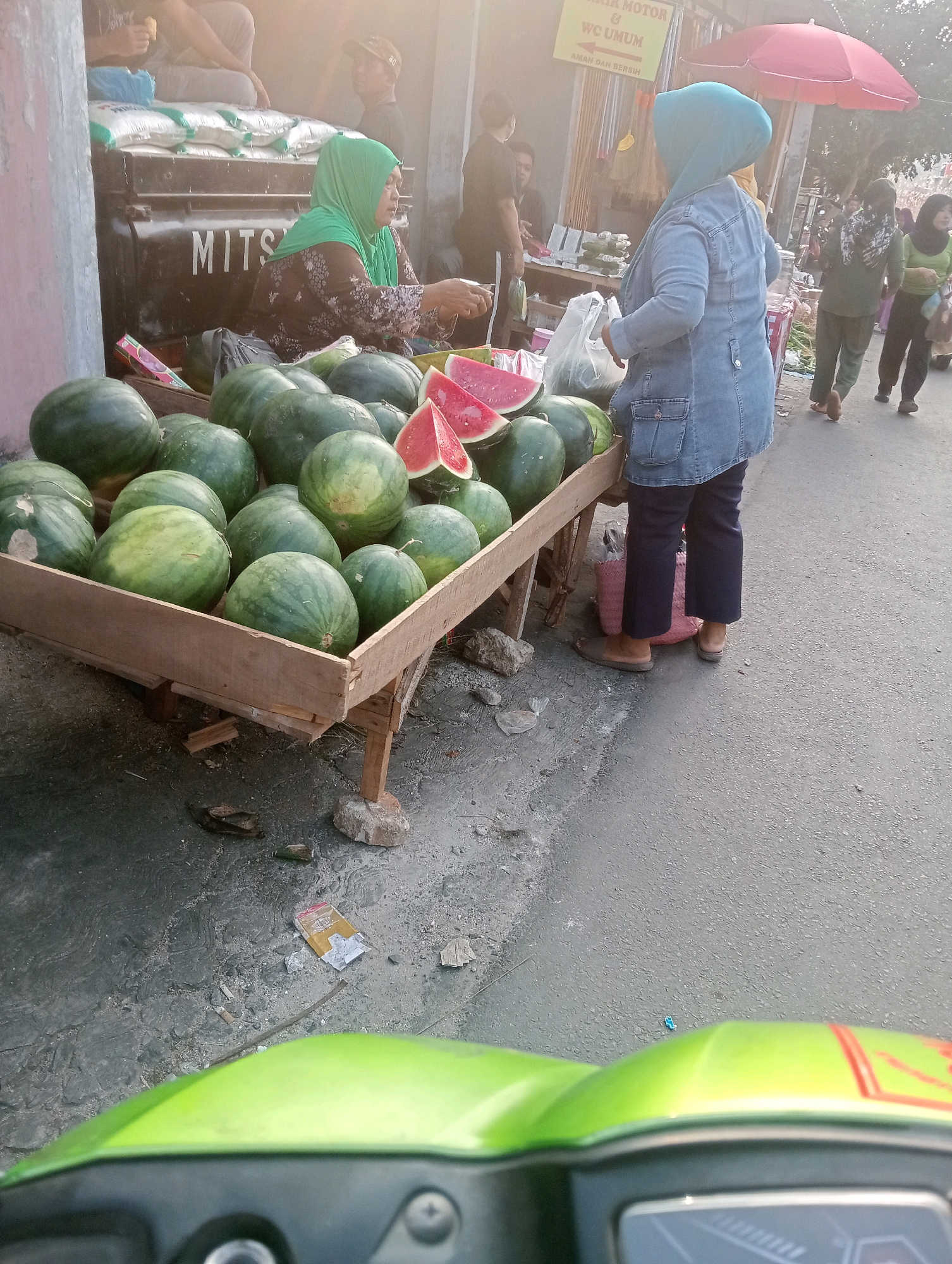 The image depicts an outdoor market scene. In the foreground, a stall is filled with numerous watermelons, some whole and some sliced. A woman wearing a green hijab and a patterned brown shirt stands behind the stall, appearing to be selling the fruit. To the right, another woman, wearing a blue denim jacket and dark pants, is facing away from the camera and also appears to be at the market. Behind them, several other people are visible in the background, with some interacting and others walking. The market seems to be set up along a paved road, with a red umbrella visible in the upper right portion of the frame. The overall atmosphere suggests a bustling marketplace.