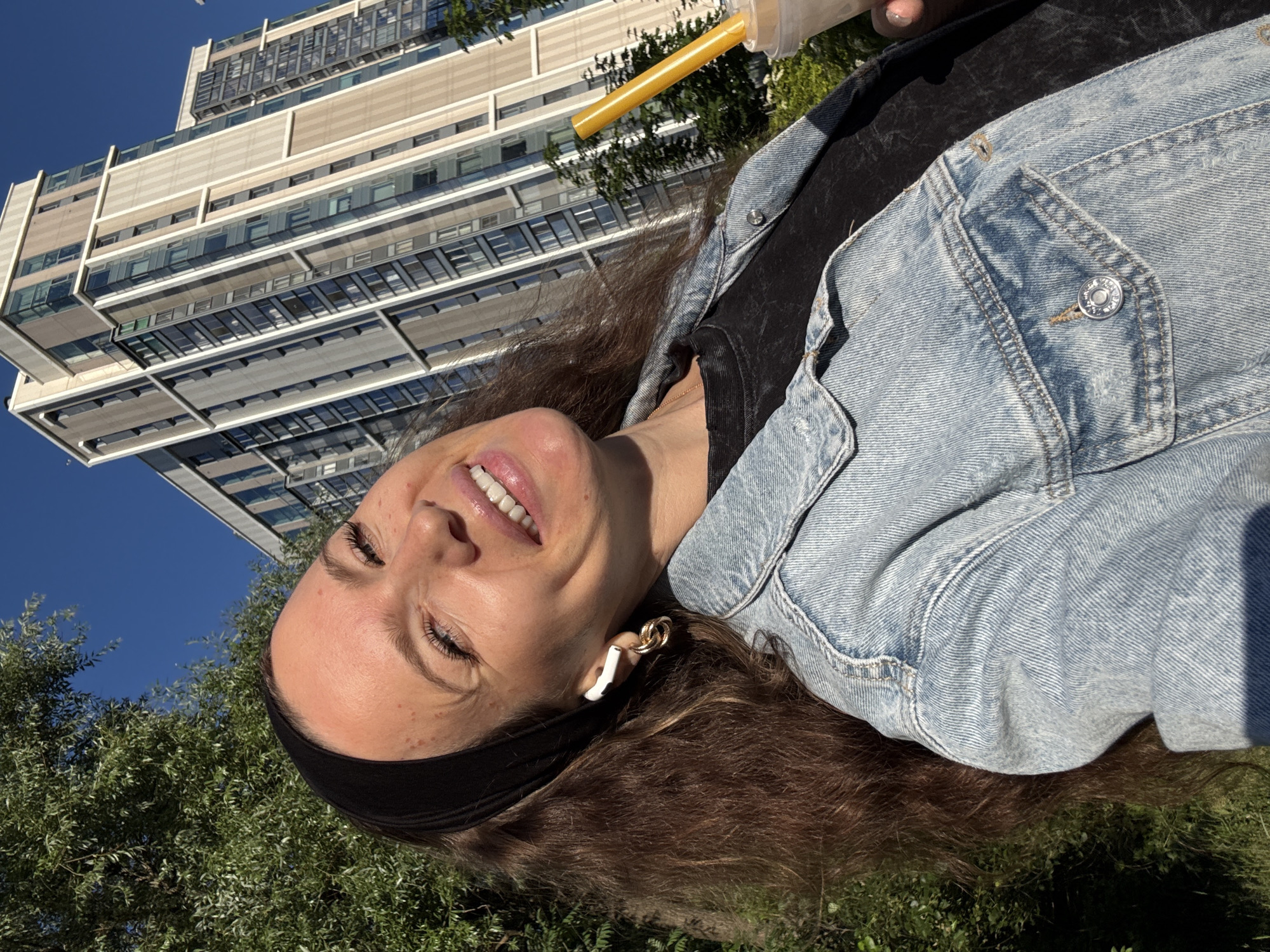 A woman is pictured outdoors in Moscow, Russia, in a selfie with a modern, tall building in the background. She is wearing a denim jacket over a black t-shirt and has a black headband on. She is smiling and holding a drink with a yellow straw. She is also wearing white wireless earbuds with gold hoop earrings. The sky is blue and the scene is lit by sunlight, indicating it is daytime. Trees and greenery are visible around her, suggesting a park or urban green space.