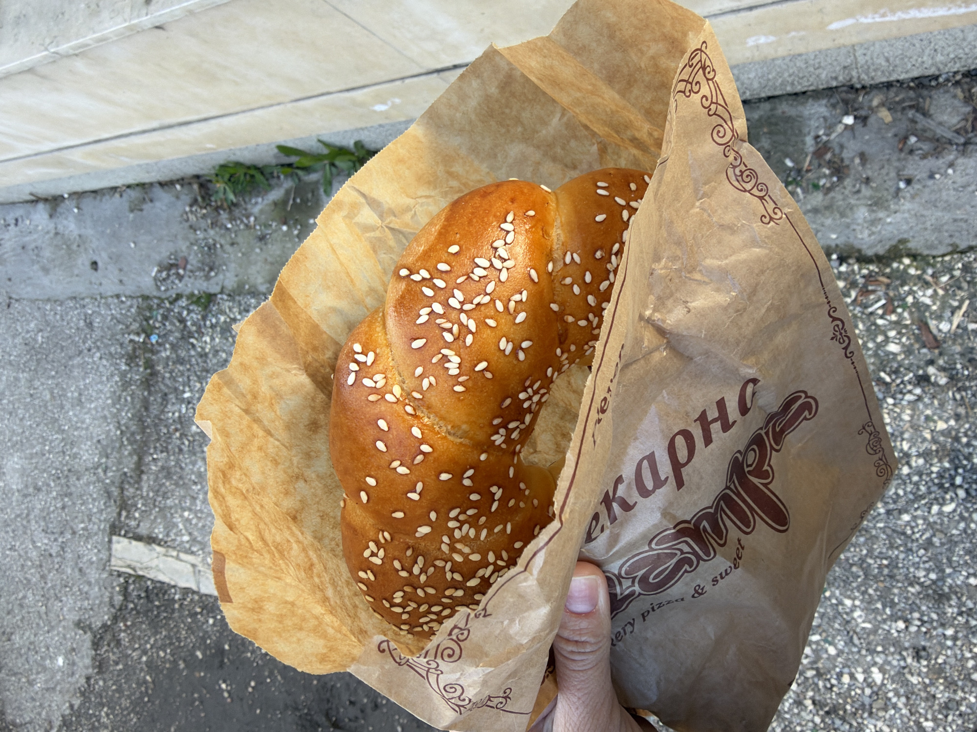 An overhead shot shows a hand holding a brown paper bag containing a sesame-seeded pretzel. The bag is printed with the word "Leкарна". The background is an outdoor scene, suggesting a city street. The location is Pleven, Bulgaria. The surface is asphalt.