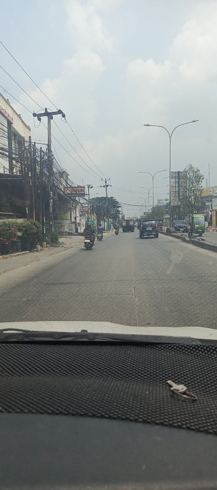 The image captures a view from inside a vehicle, looking out at a moderately busy street in Sukaresmi, Indonesia. The perspective is from the driver's seat, showing a portion of the dashboard with a textured surface in the foreground. The road ahead is paved and appears to be a main thoroughfare, with multiple lanes of traffic.
Several vehicles are visible: a white pickup truck in the distance on the right side of the road, a black SUV in the center lane, and a green three-wheeled vehicle (possibly an auto-rickshaw or "bajaj") parked on the right side. Further down the road, a truck is in the left lane. Several motorcycles are also present, with riders wearing helmets and casual clothing.
On the left side of the street, there is a row of buildings. The closest building has a light-colored facade and is adorned with numerous electrical wires and utility poles, suggesting a densely wired urban area. A sign that reads "JOTUN" is visible on one of these buildings. Further down the street, there are more buildings, some with colorful banners and flags, and trees. On the right side of the road, there are some construction materials and trees. The sky is overcast with some visible clouds. The overall impression is that of a typical day in a developing urban environment in Indonesia.