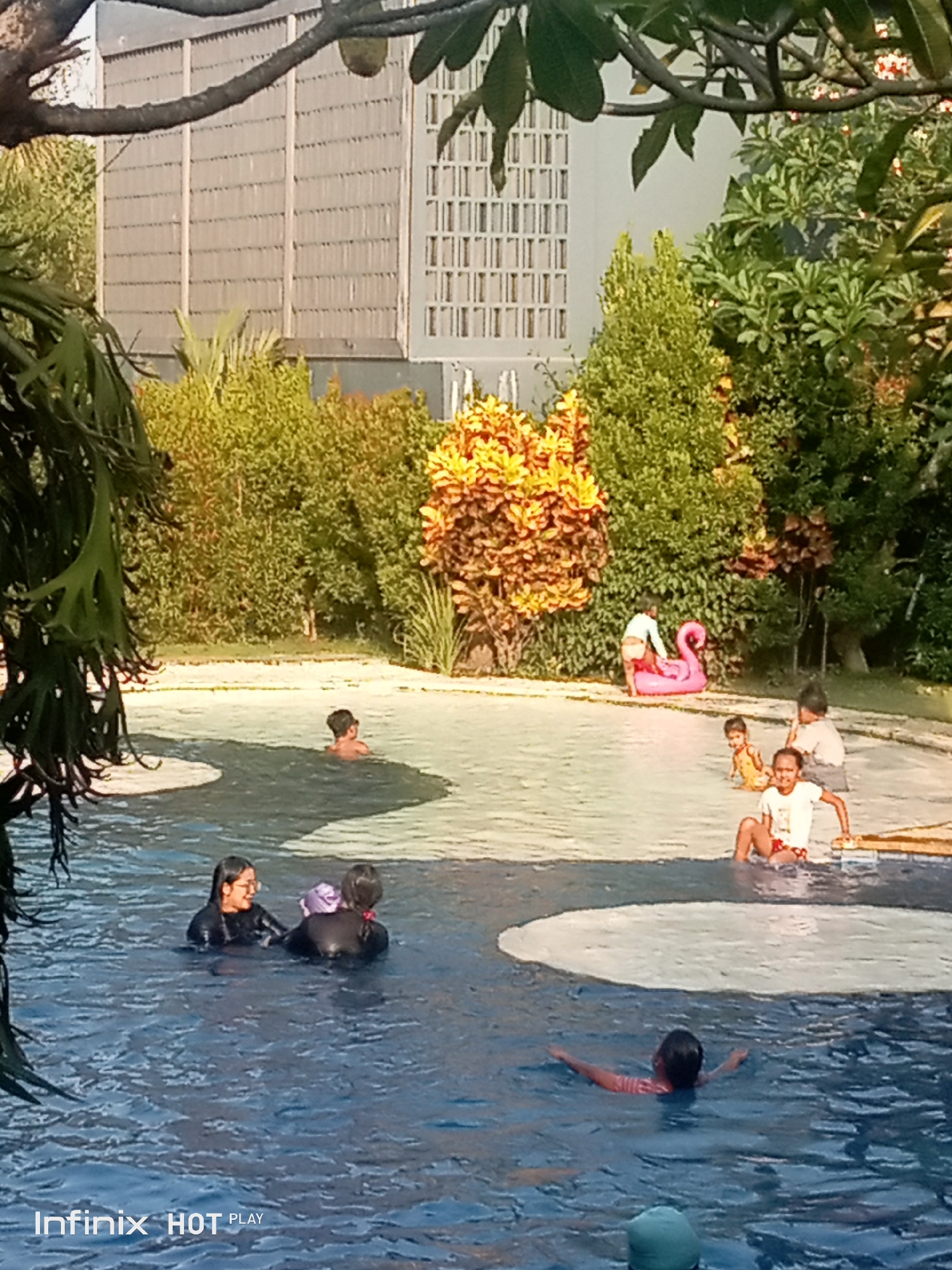 A group of children and adults are at a swimming pool. In the foreground, two adults are swimming, and one child is also in the pool. In the background, several children are playing near the pool's edge. One child is sitting by a pink inflatable flamingo, and another child is standing next to it. The pool is surrounded by lush greenery and a building with a distinctive facade. The scene appears to be in a tropical location. The Infinix HOT PLAY watermark is visible in the bottom left corner of the image. The image is likely from Ubud District, Indonesia, given the architectural style and vegetation.