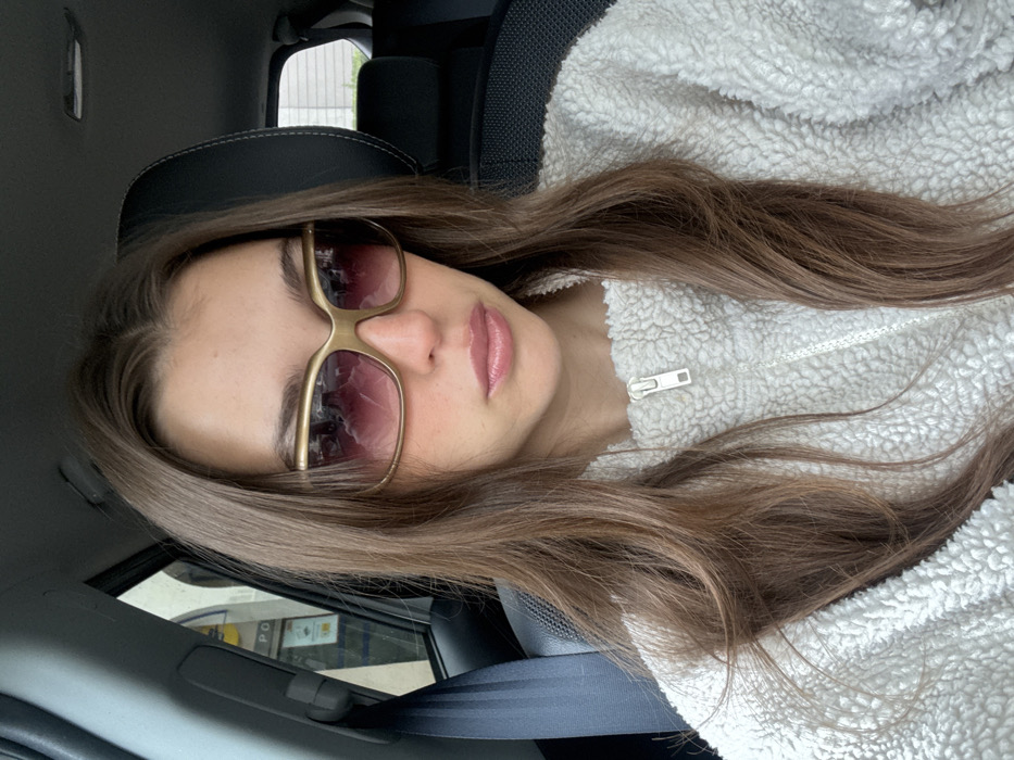 A young female is depicted seated in the front passenger seat of a vehicle in Loire-Authion, France. She has long brown hair, parted centrally, and is wearing a cream-colored, textured quarter-zip fleece pullover with a white zipper. Her eyes are obscured by large, rectangular sunglasses featuring gold-toned frames and gradient purple-tinted lenses. Her lips are closed and coated with a light pink gloss. A black vehicle seatbelt is positioned across her left shoulder and chest. Through the vehicle's left-side window, a blurry exterior shows a building with a yellow commercial sign, possibly from a LIDL supermarket, displaying indistinct text and partial numbers (e.g., '3.90'). A black vehicle headrest is visible behind her, and blurred green foliage is discernible through the rear passenger window.