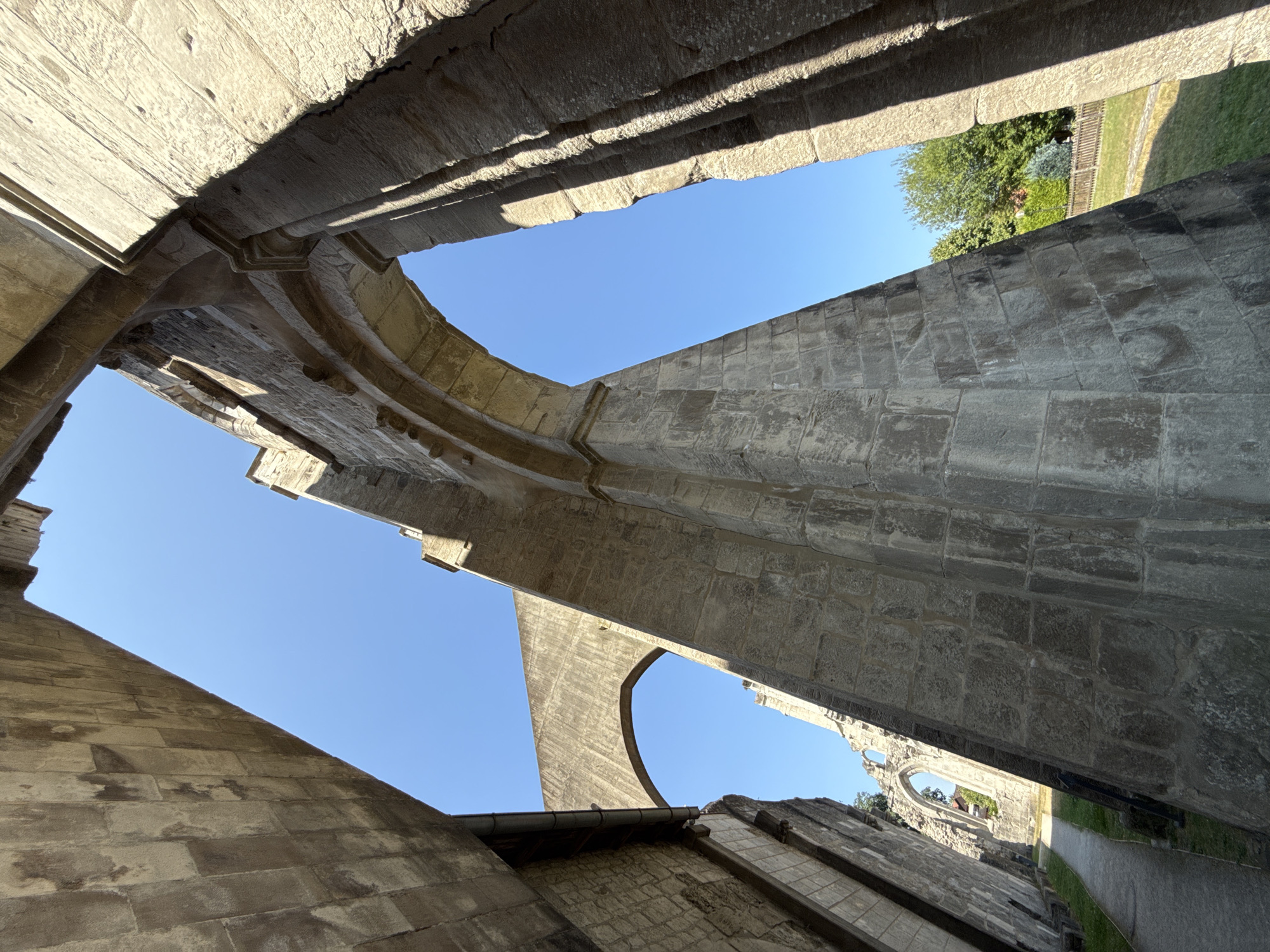 The image captures the ruins of the Walkenried Abbey in Walkenried, Germany. The viewpoint is looking upwards through massive stone arches and walls, showcasing the architectural remnants of the historical abbey. The sky is a clear, bright blue. The stone structures are weathered and show signs of age, with visible masonry and varying textures. In the distance, glimpses of green trees, a wooden fence, and some modern buildings are visible, indicating the abbey is situated within or adjacent to a more contemporary environment. A paved pathway can be seen leading through the ruins. There are no people present in the image.