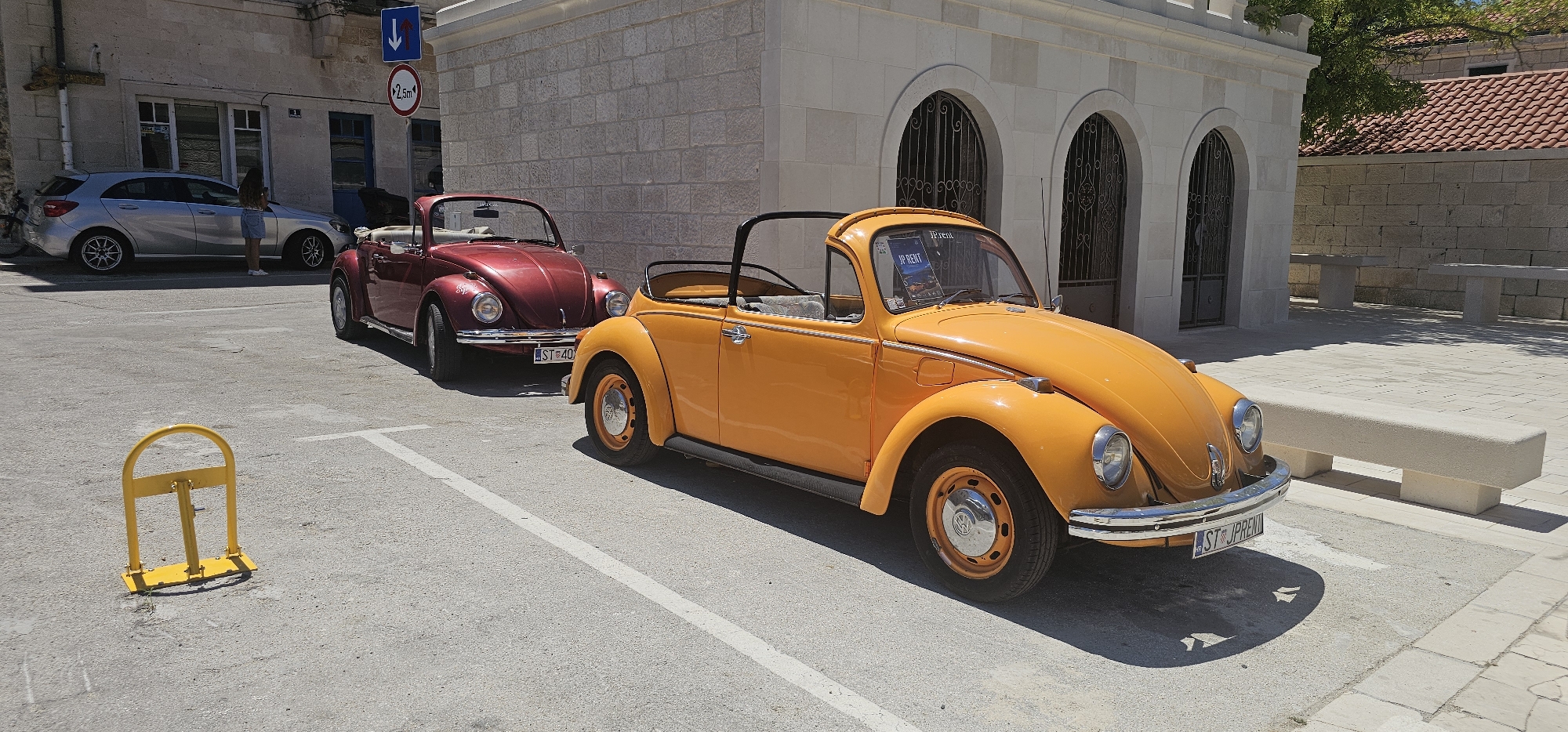 Two classic Volkswagen Beetle convertibles, one maroon and one bright orange, are parked side-by-side on a paved surface in what appears to be a town square or parking area in Općina Pučišća, Croatia. The maroon Beetle is parked behind the orange one, slightly angled to the left. The orange Beetle is prominently positioned in the foreground, facing to the right, with its top down. A parking barrier, painted bright yellow, is visible in the lower left quadrant of the image. In the background, a stone building with arched entrances and a tiled roof is visible, suggesting a Mediterranean architectural style. A silver hatchback car is parked further back, to the left of the Beetles, with a person standing beside it. The ground is paved with concrete or asphalt, and white parking lines are marked on the surface. The scene is bathed in bright sunlight, casting shadows from the vehicles and a bench in the background. The overall impression is a sunny day in a picturesque European town.