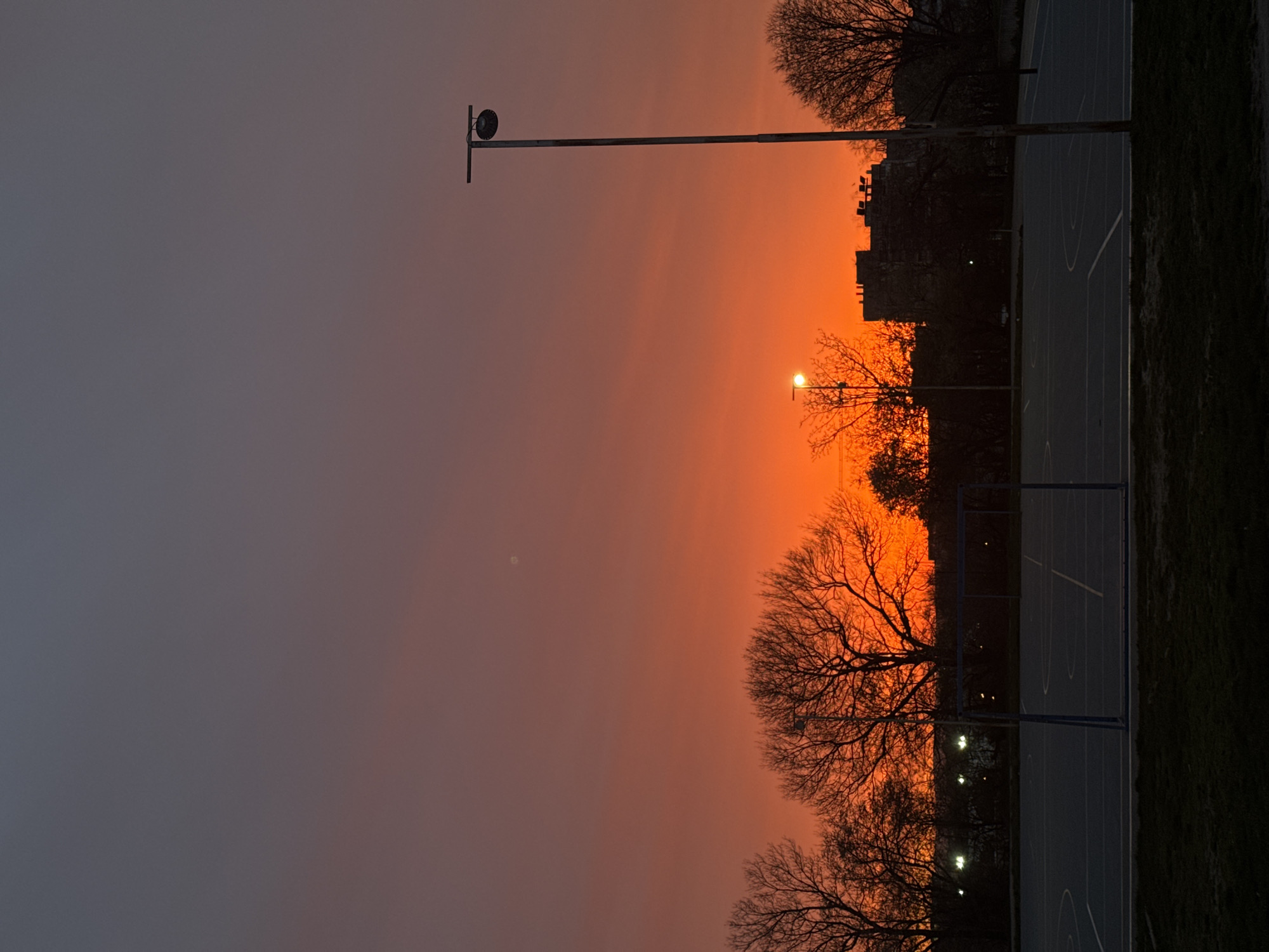 The image shows a dusky scene in what appears to be Varna, Bulgaria, given the architectural style and the presence of a sports court. The sky is dominated by gradient hues ranging from grey at the top left to orange closer to the horizon.
There are silhouettes of trees, and buildings against the fiery sky, suggesting twilight. A tall streetlight with a speaker is visible on the left side of the frame. A sports court is to the right, and appears to be a basketball court with visible markings.