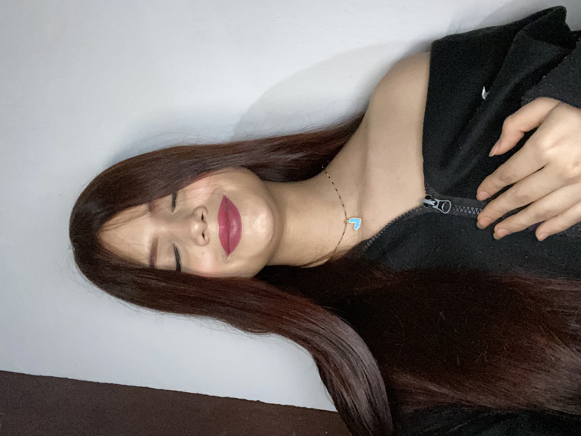 The image shows a woman with long, straight, reddish-brown hair lying on a surface. She is wearing a black zip-up jacket and a necklace with a light blue heart pendant. Her eyes are closed, and she is wearing dark red lipstick. The background appears to be a white wall. There is no indication of a specific location such as Juan Griego, Venezuela.